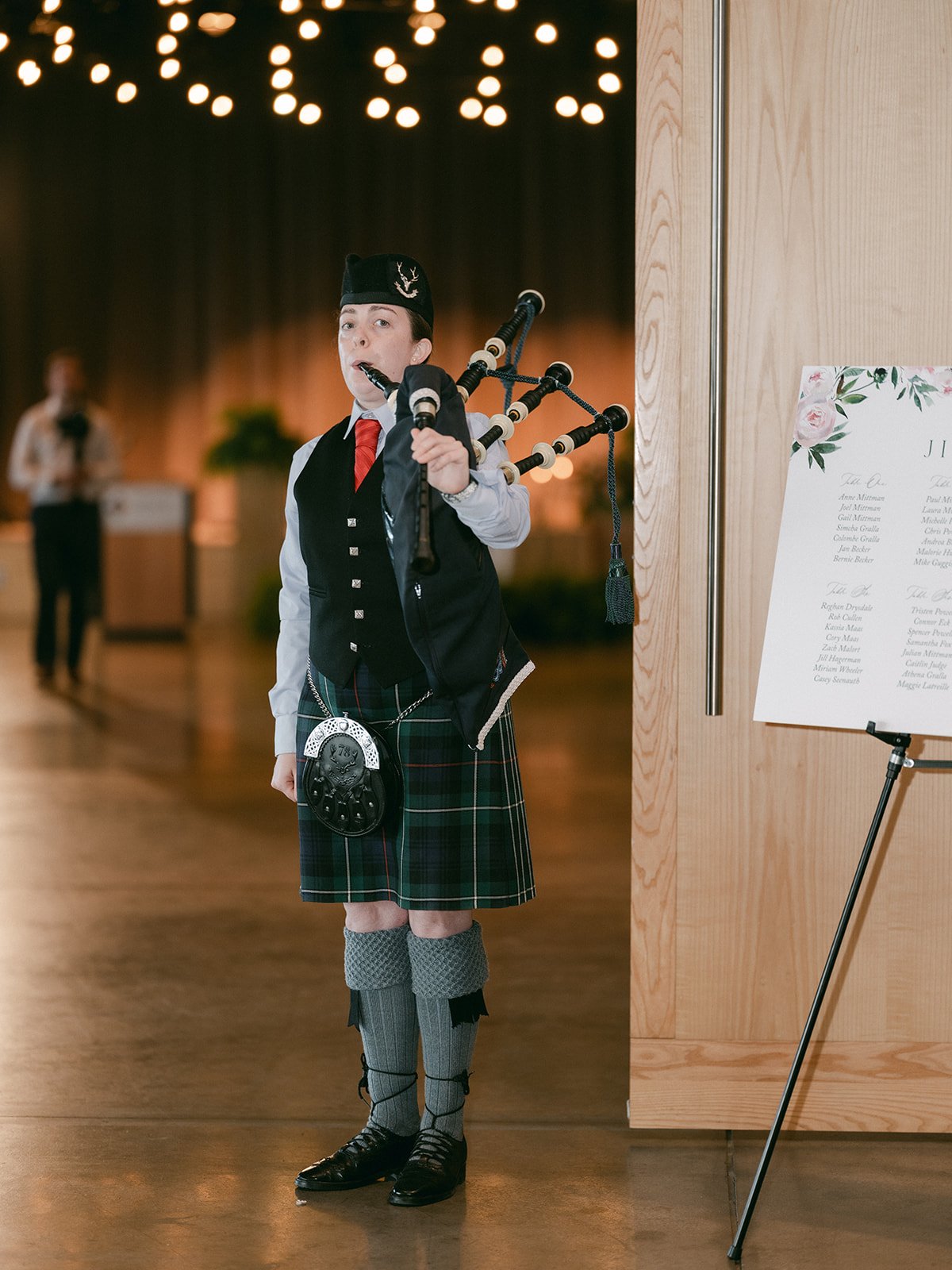 Person wearing traditional Scottish attire playing bagpipes indoors with decorative lights and a sign on the side.