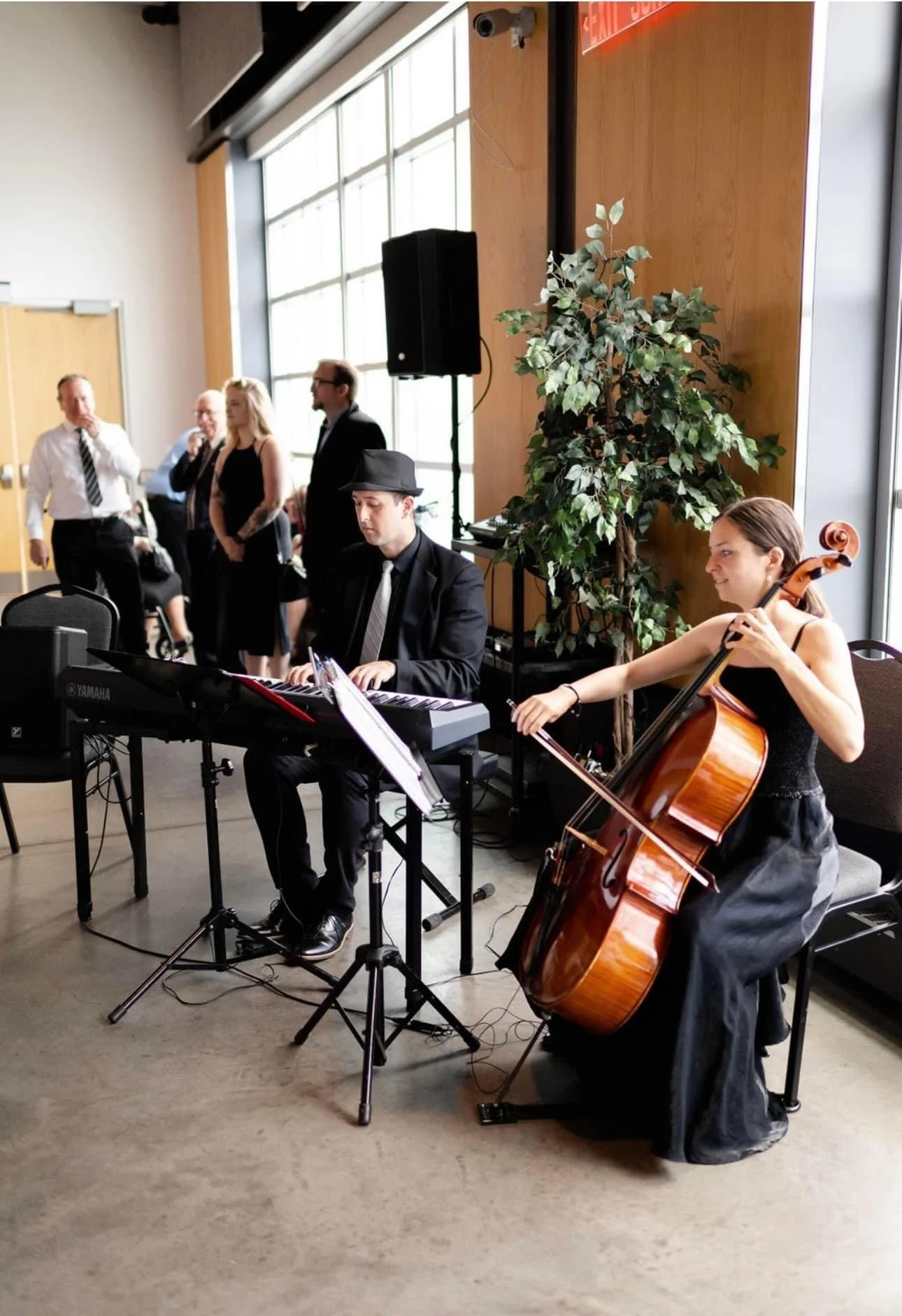 Musicians at an event with a pianist and cellist performing in front of standing audience in a modern venue with a large window and plant decor.