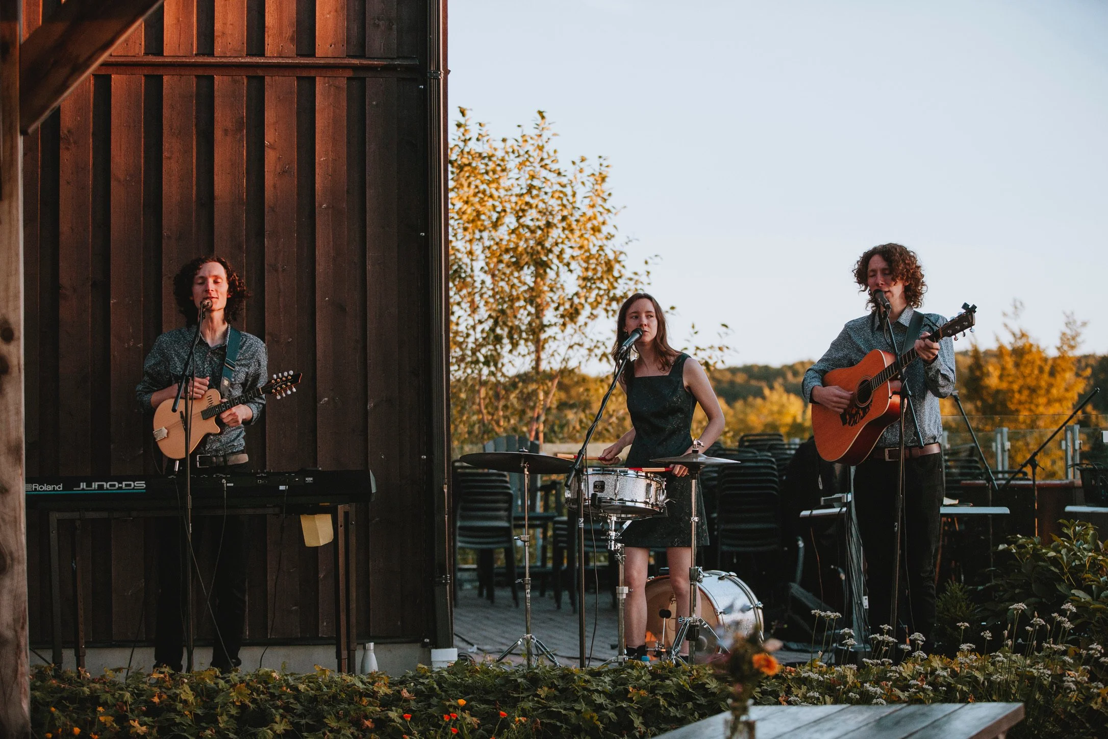 Three musicians playing outdoor music, with keyboard, drums, and acoustic guitar, against a wooden wall backdrop during sunset.