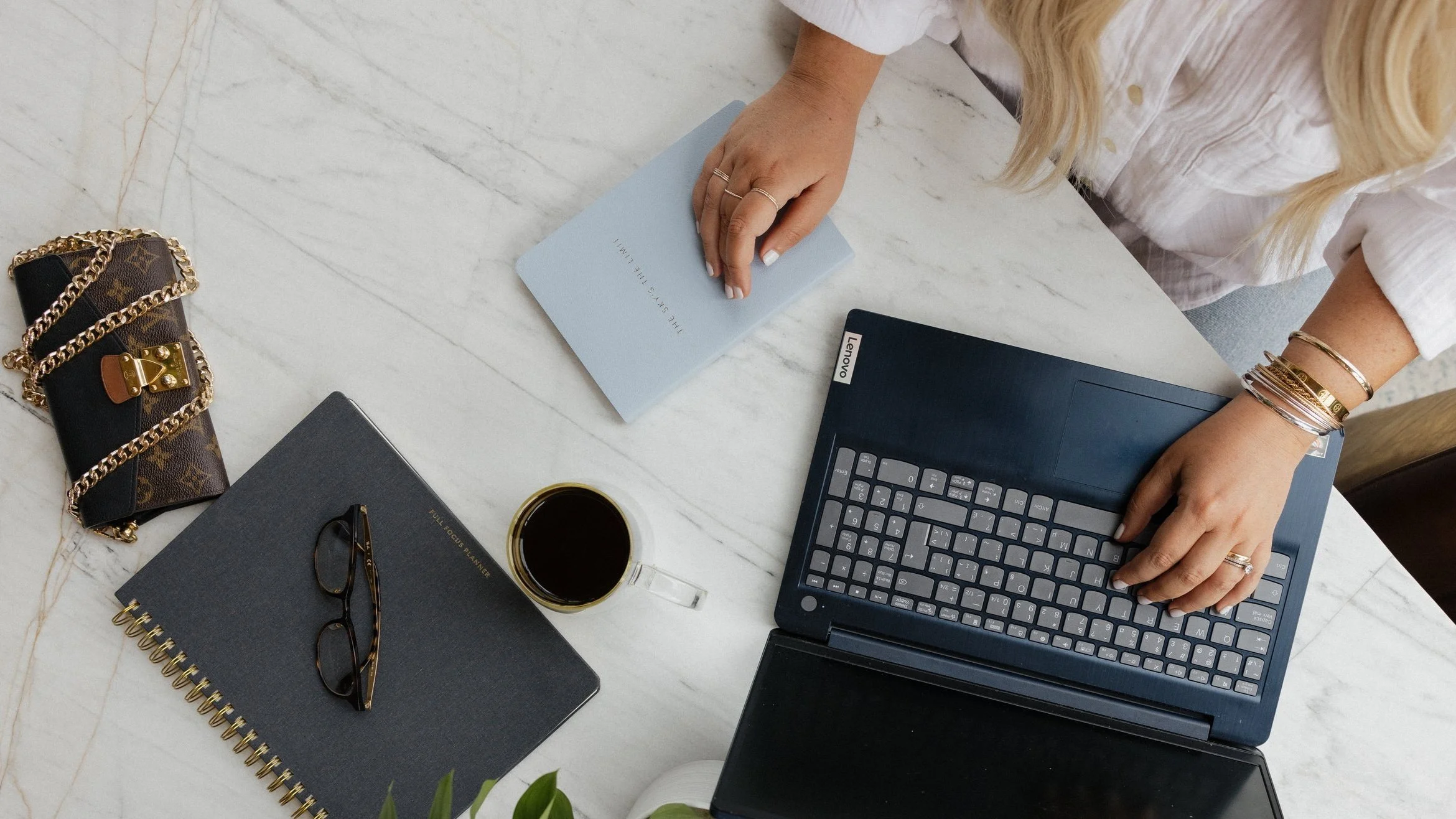 Woman's hands working on a Lenovo laptop with a notebook, coffee cup, glasses, and designer purse on a marble desk.