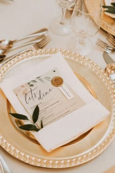Elegant dinner table setting with a gold beaded charger plate, white linen napkin, and an ornate menu card. Wine glasses and silverware are neatly arranged beside the plate.