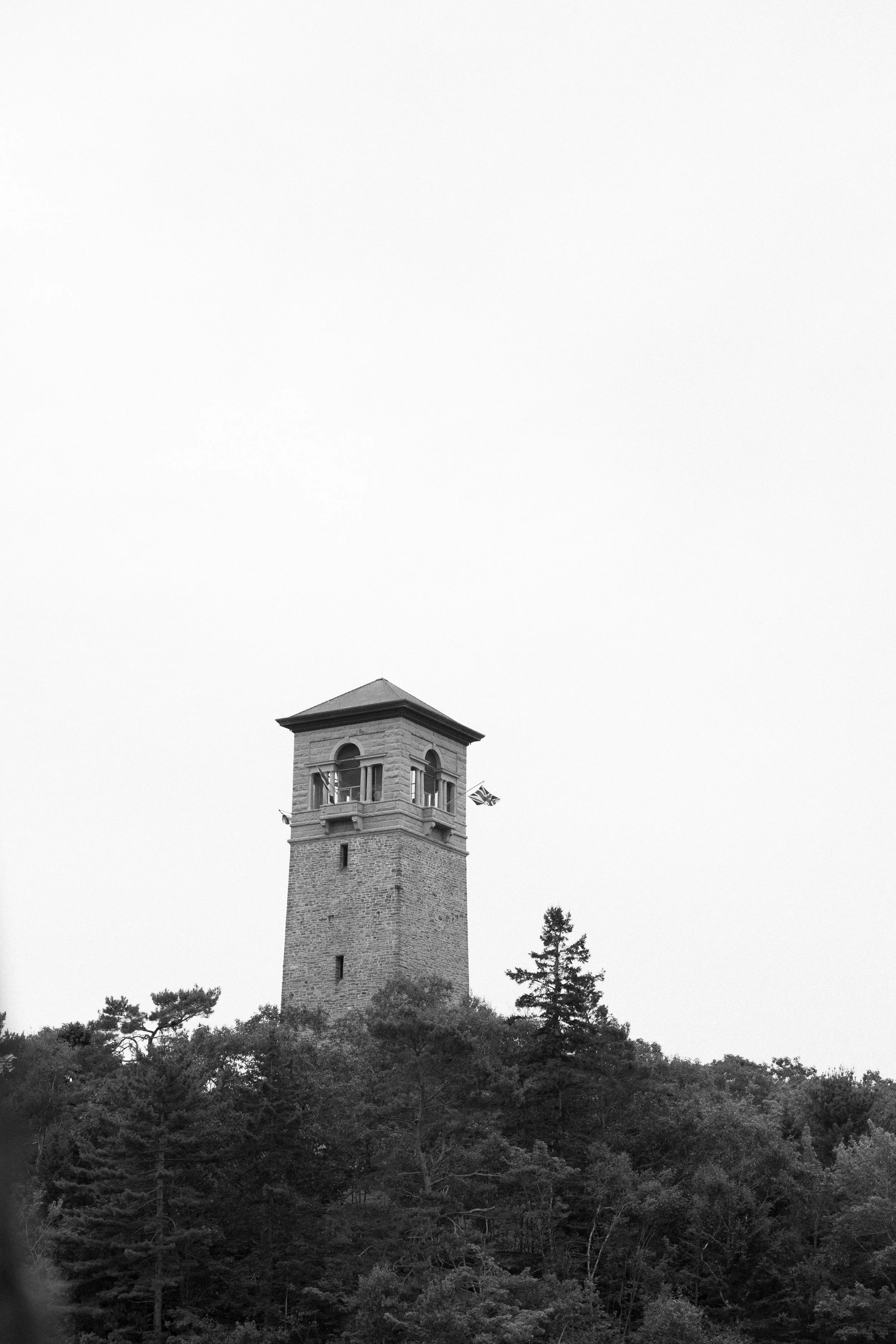 Black and white photo of a tall stone tower with a pointed roof, surrounded by trees.