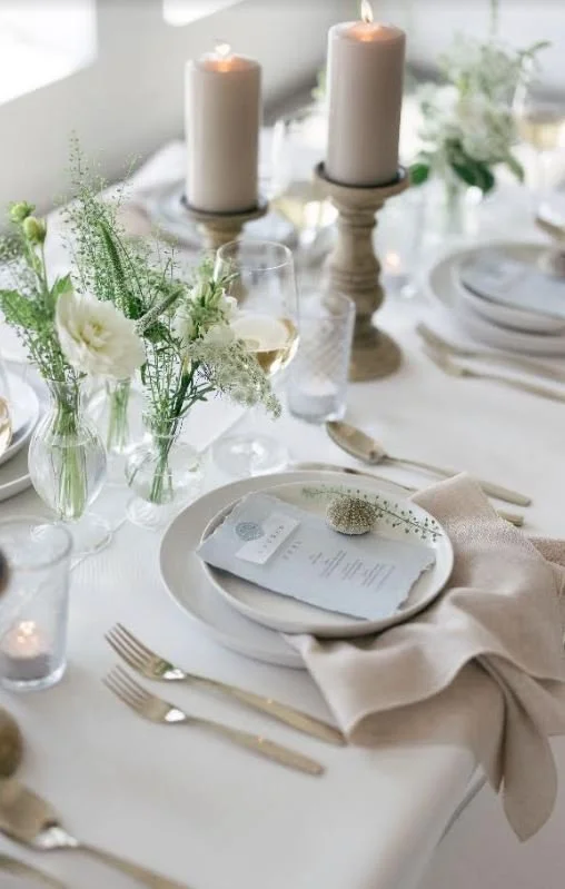 Elegant table setting with white plates, silverware, beige napkins, menu cards, candles, and floral arrangements in small vases.