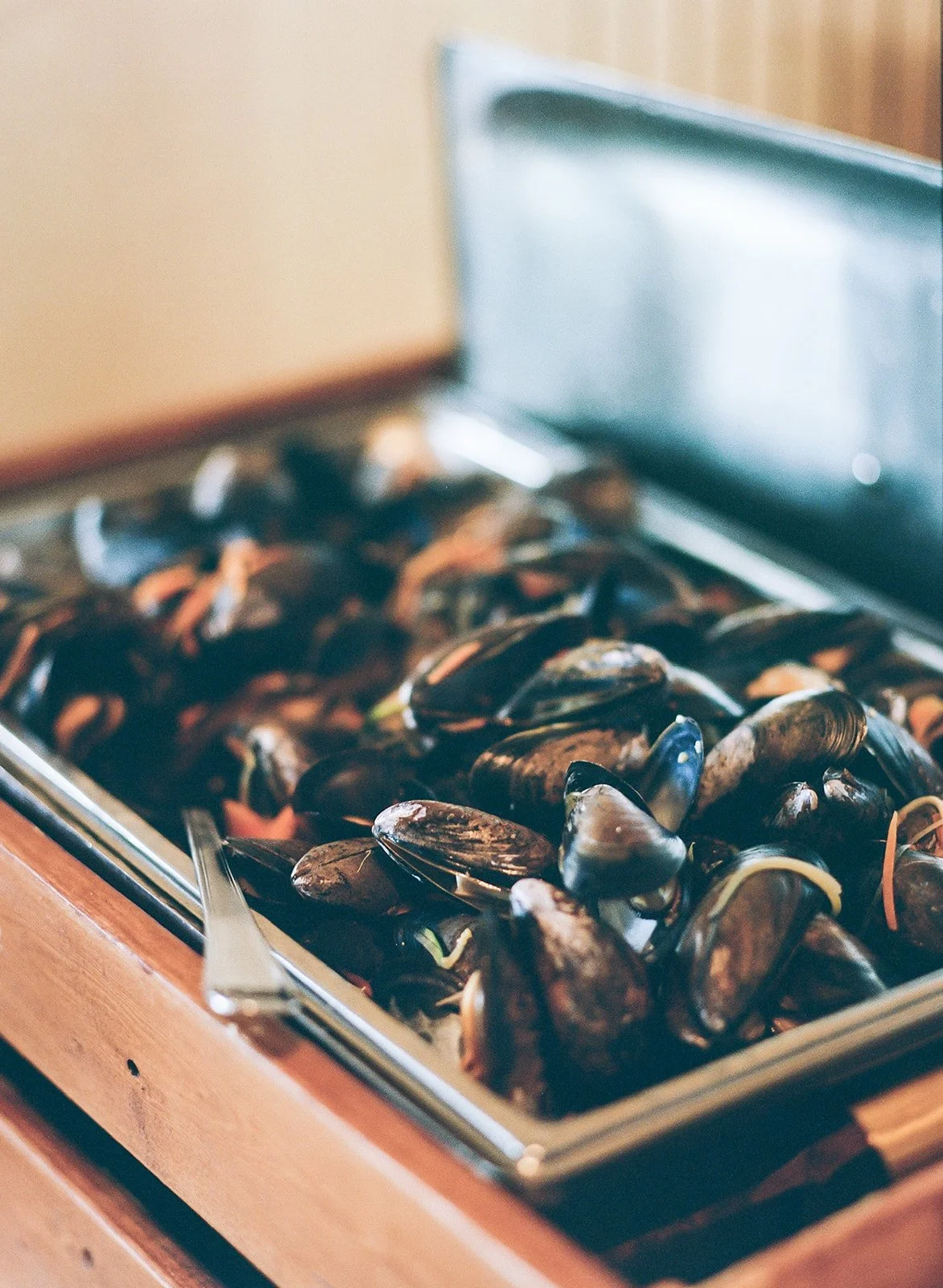 A stainless steel tray filled with cooked mussels, placed on a wooden surface, with a serving spoon on the side.