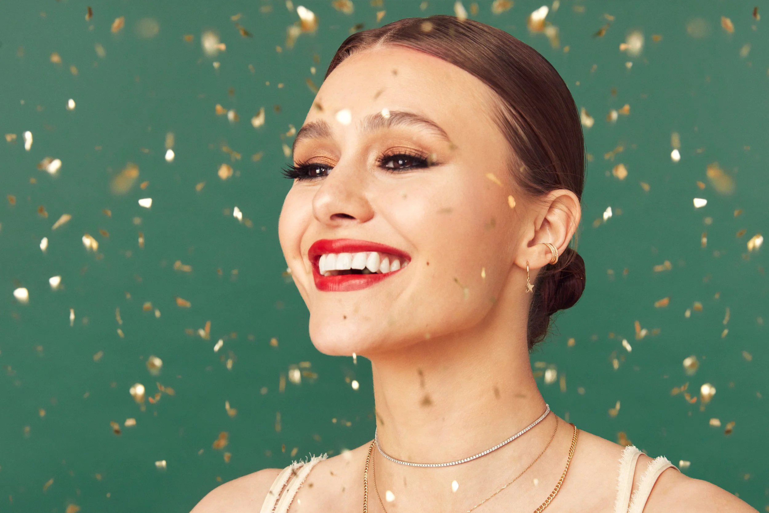 A young woman with red hair styled in a bun, smiling with red lipstick, surrounded by gold confetti against a green background.