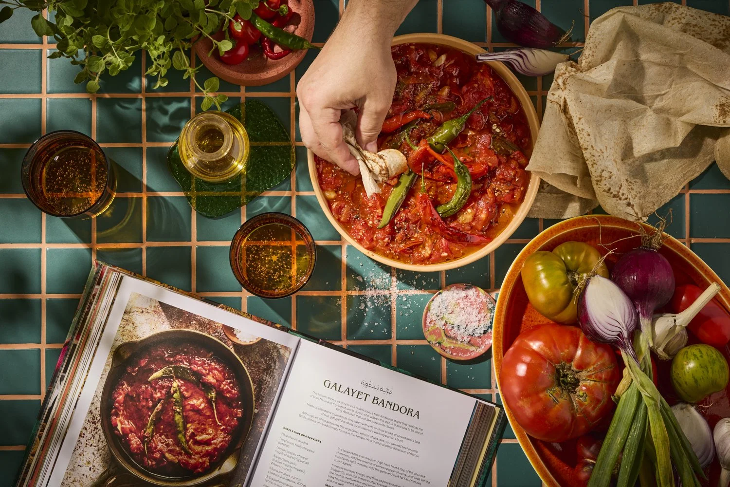 A person preparing a dish with chopped vegetables, chili peppers, and a tomato-based mixture in a bowl on a tiled table. There are glasses of beverage, a cookbook open to a tomato stew recipe, a bowl of fresh tomatoes, onions, garlic, and herbs, and 
