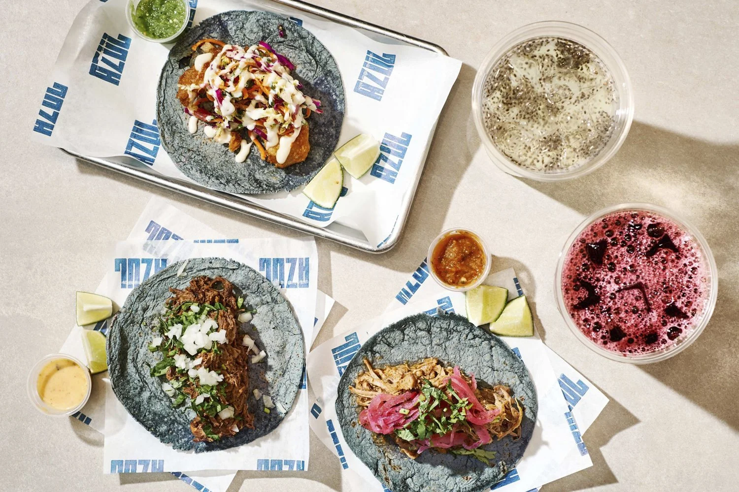 Three tacos on black plates with lime wedges, two cups with sour green sauce and pink drink, and a glass of clear soda with ice on a beige table.