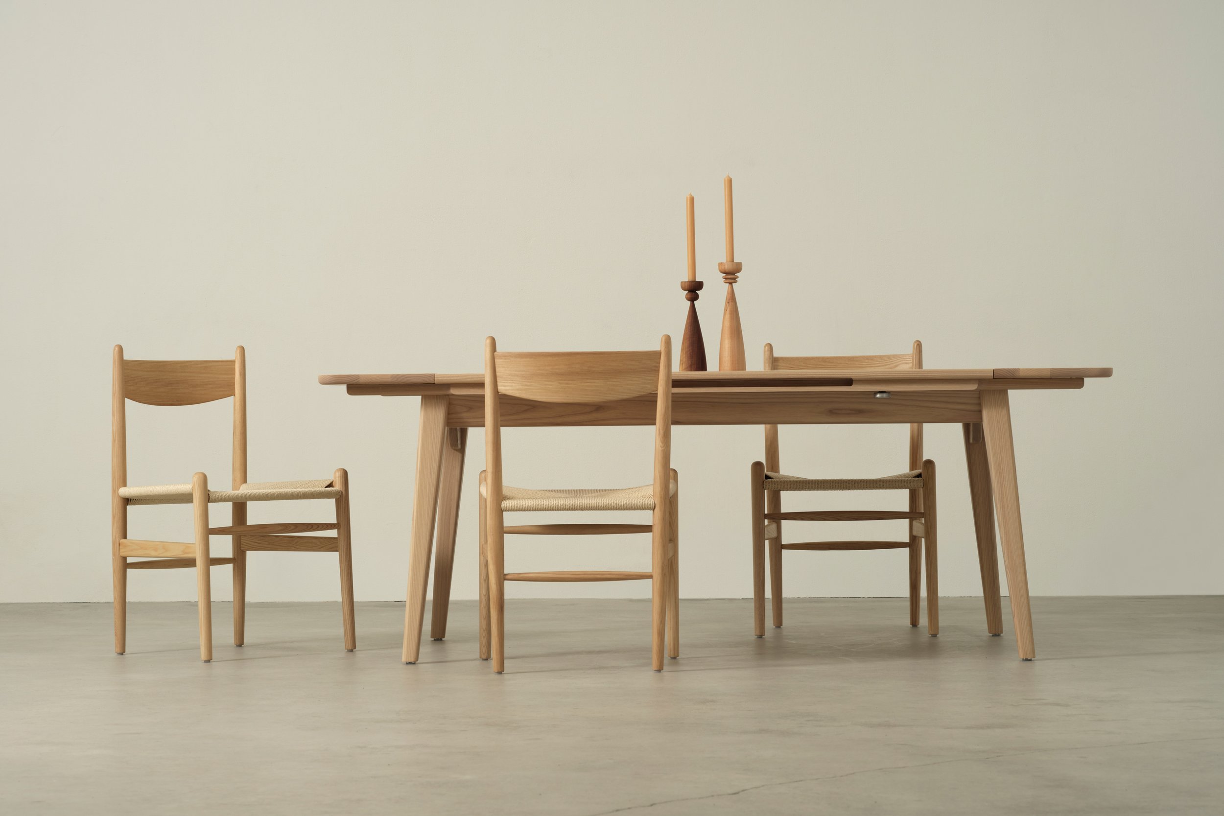 Minimalist dining table with three chairs and two candlesticks on a plain wall background.