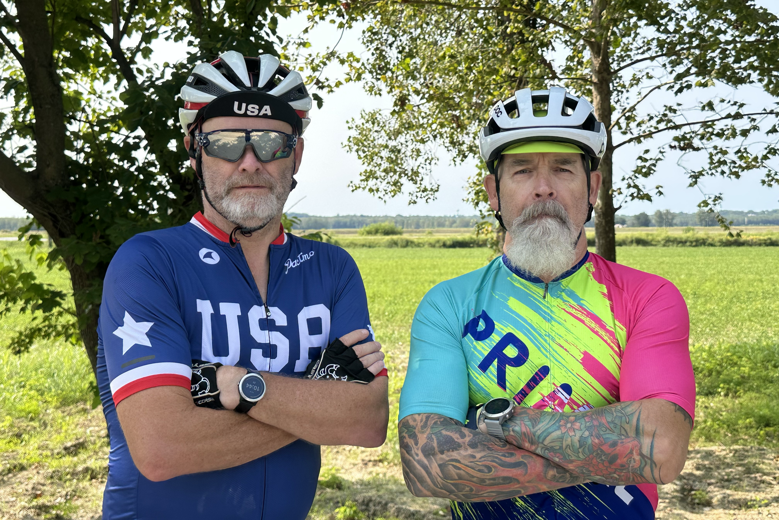 Two men standing outdoors under a tree with arms crossed. Both are wearing cycling helmets, sports sunglasses, and colorful cycling jerseys. The man on the left wears a blue USA jersey, a white and red helmet, and sunglasses. The man on the right wea