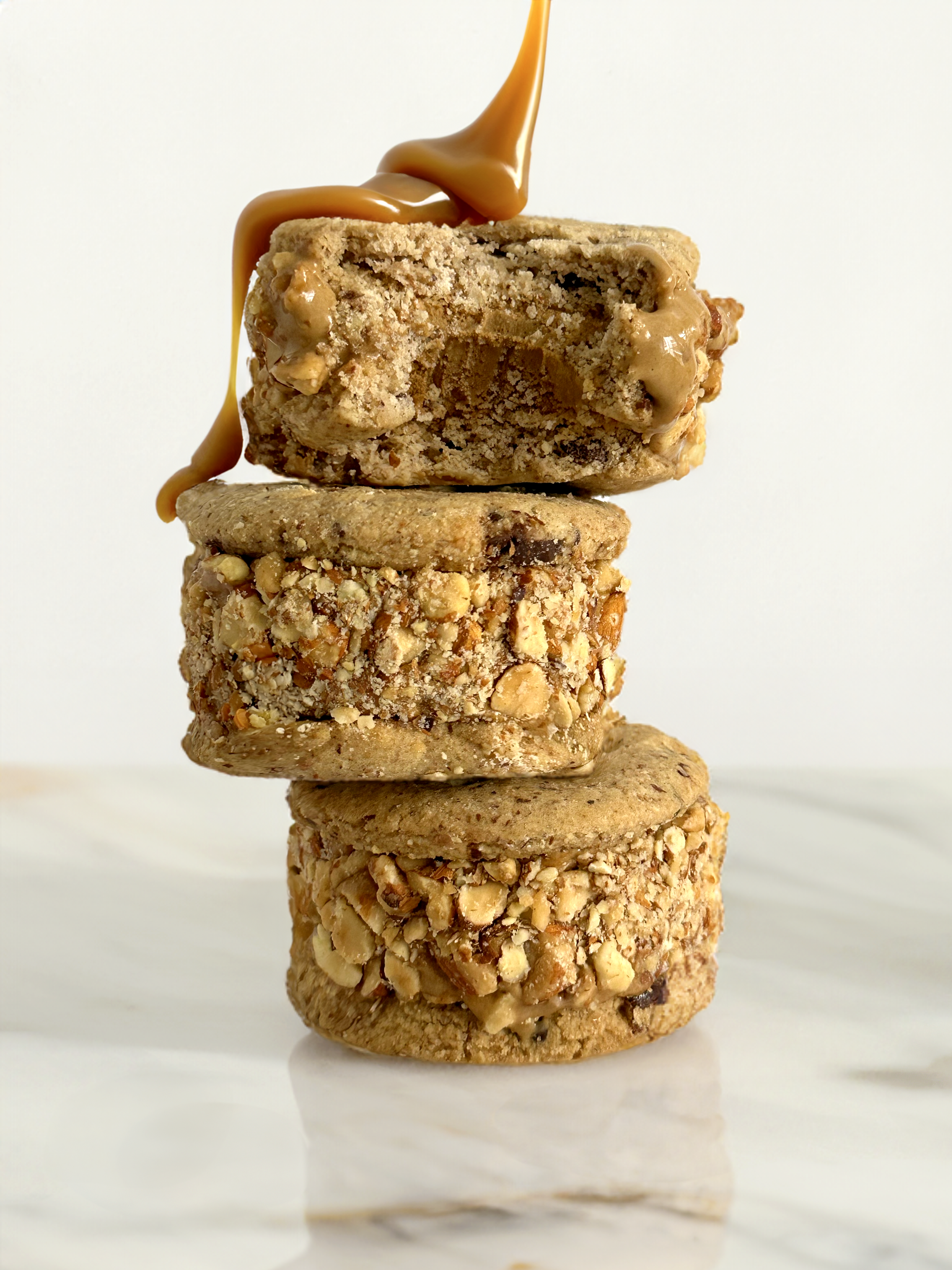 Stack of three cookies with caramel drizzle on top, including oatmeal, peanut butter, and chocolate chip cookies, on a white surface.