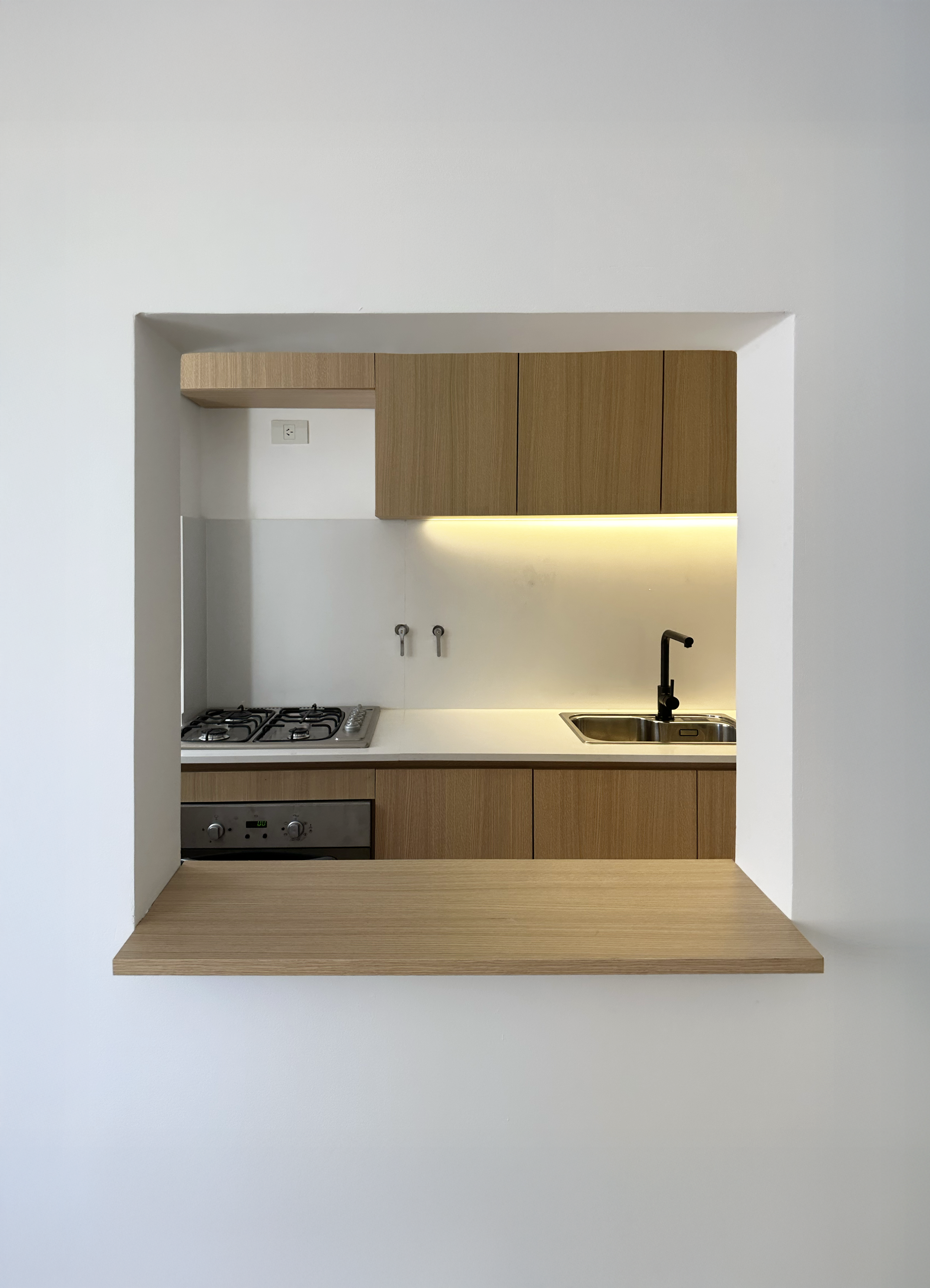 The image shows a view through a rectangular opening in a white wall into a modern kitchen with wooden cabinets, a sink with a black faucet, a stovetop, and a built-in oven.