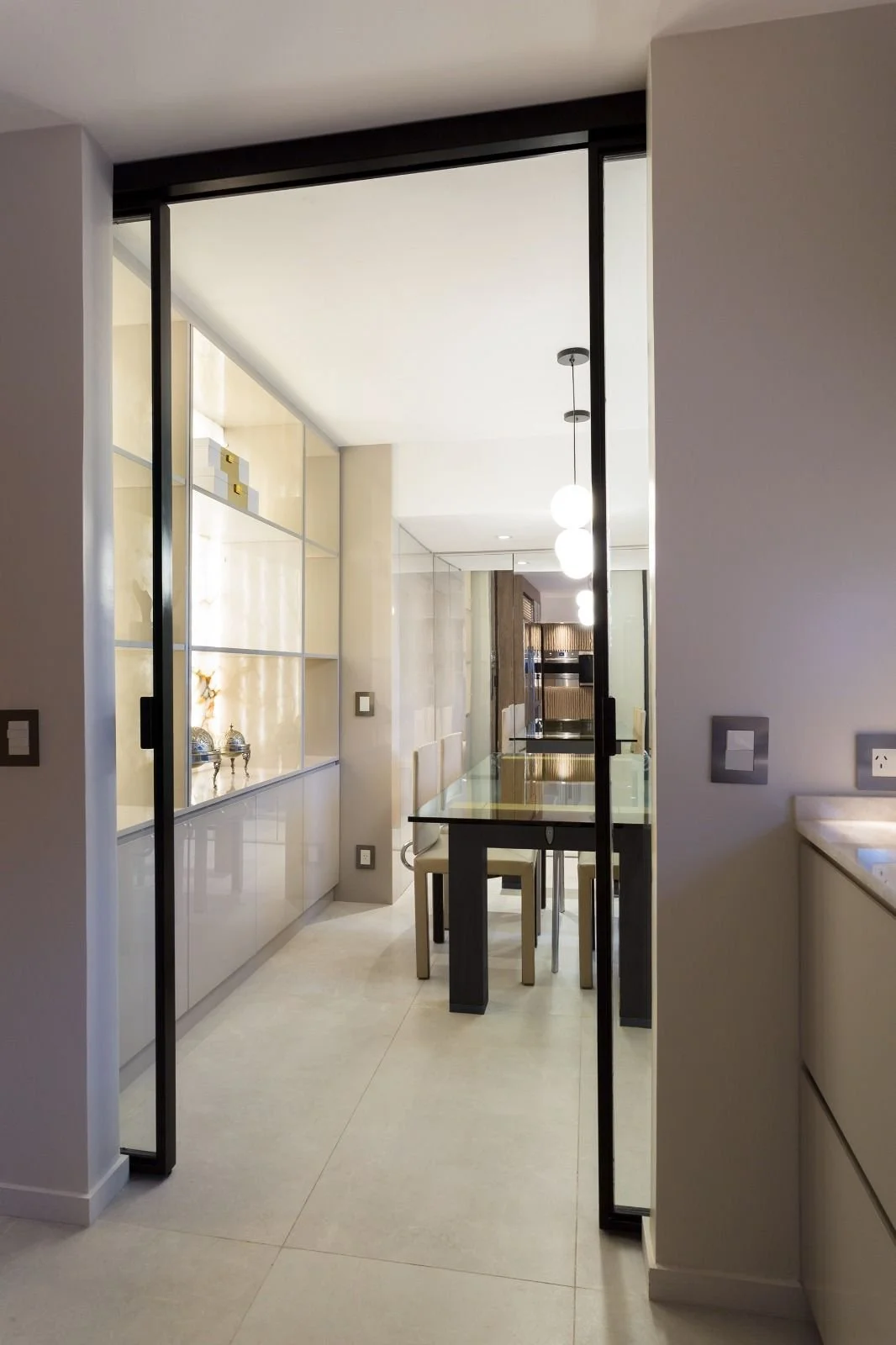 Modern dining area with a glass-top table, upholstered chairs, pendant lighting, and a built-in cabinet with decorative items, separated by black-framed sliding glass doors.
