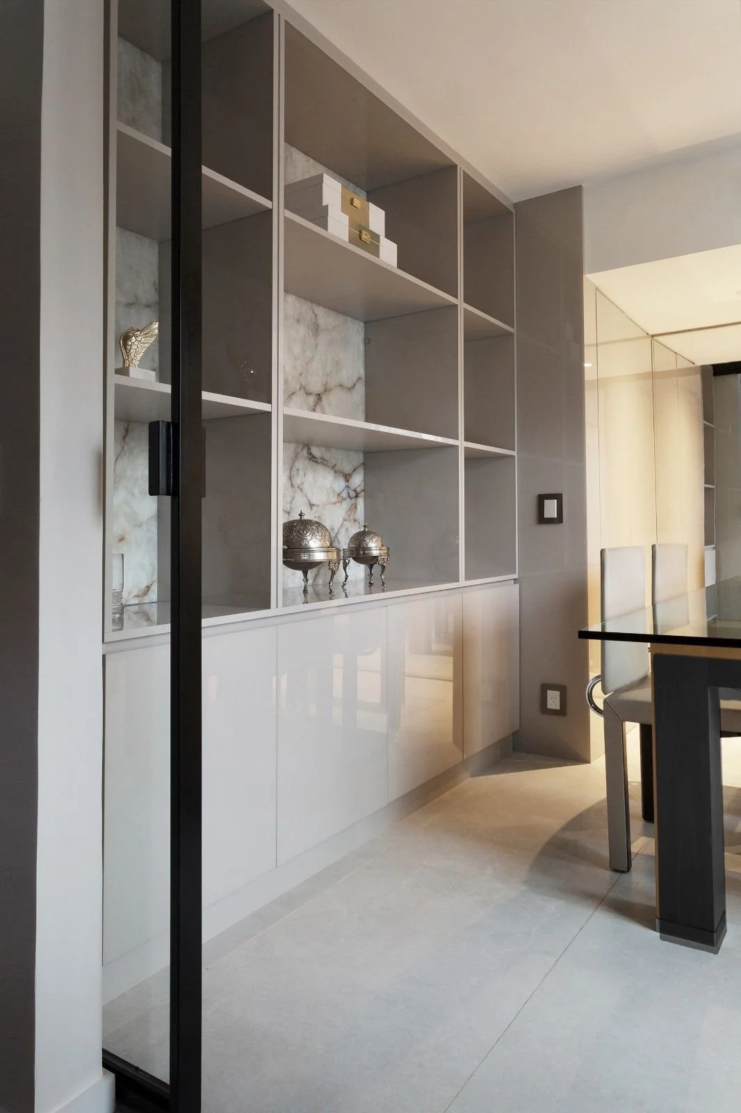 Modern built-in shelves with decorative silver box containers on marble background, beige flooring, and part of dining chairs in a contemporary interior.
