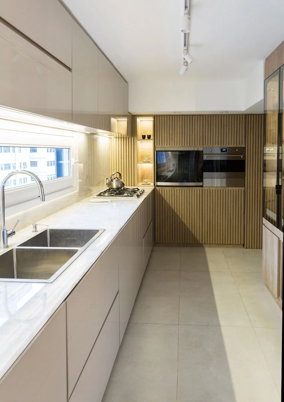 Modern kitchen with white counters, built-in oven, microwave, sink, and wooden cabinetry, illuminated by track lighting and natural light from a window.