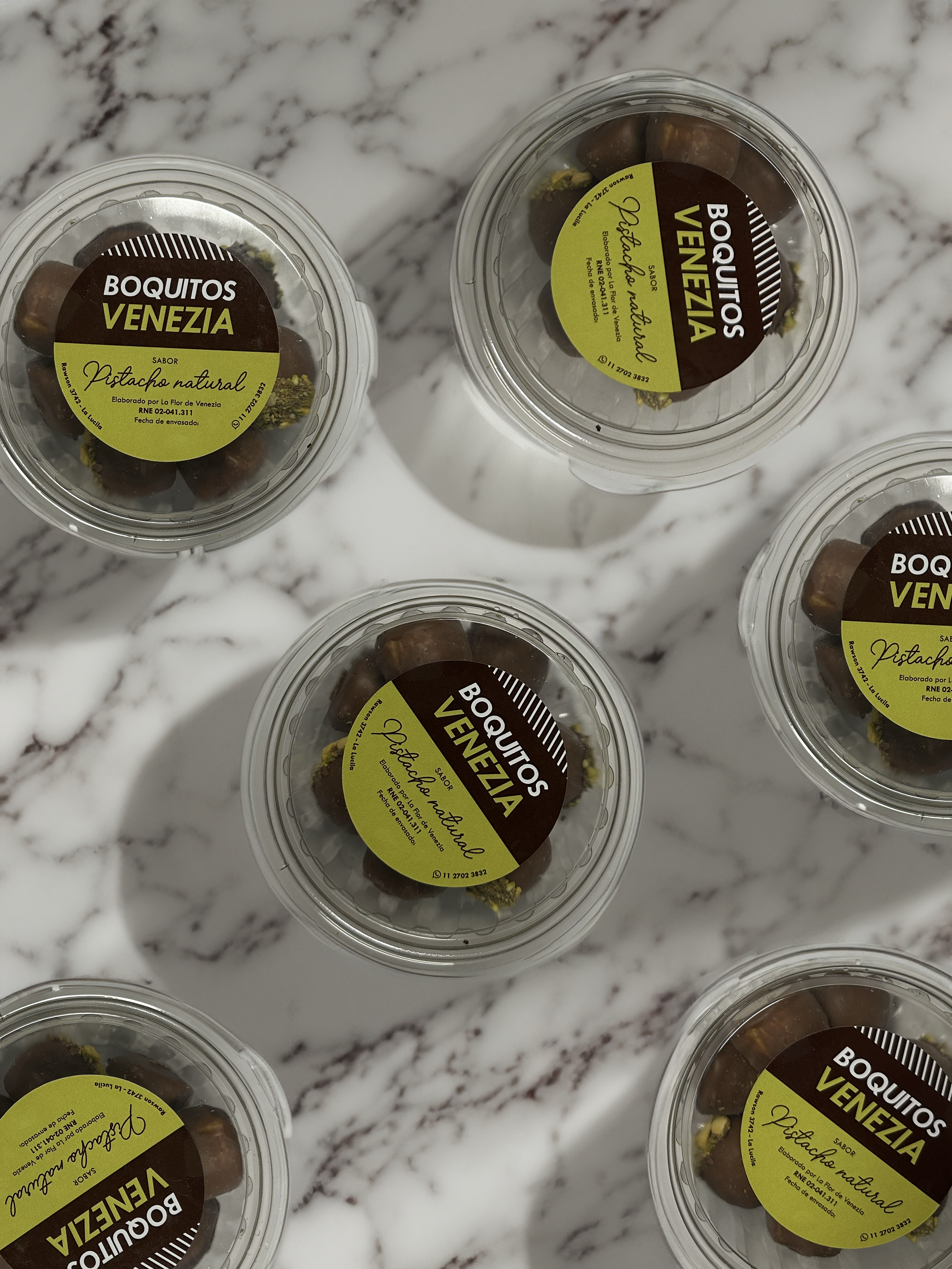 Multiple containers of pistachio nuts labeled 'Boquitas Venezia' on a marble surface.