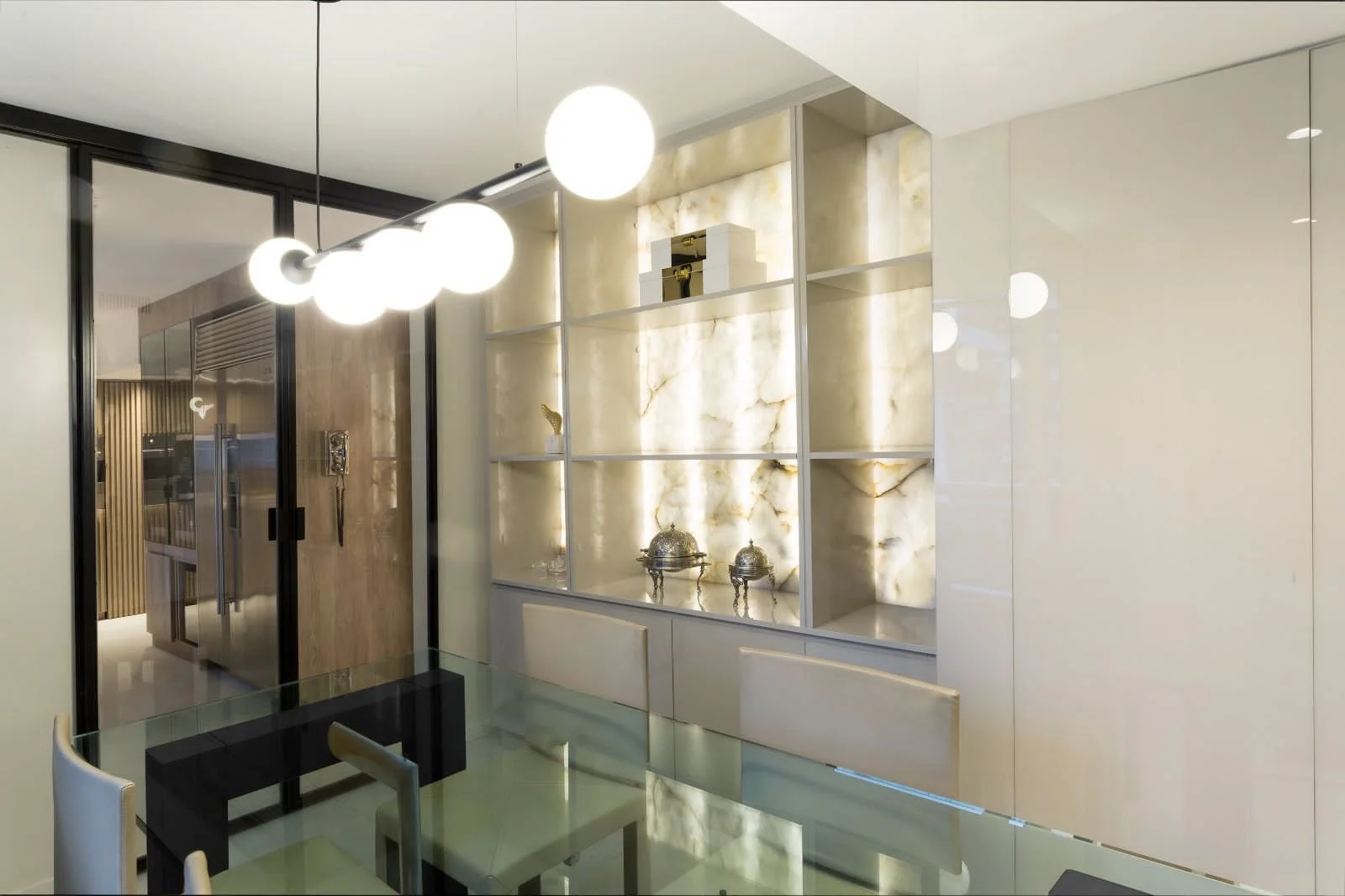 Modern dining area with glass table, beige chairs, illuminated built-in shelf with onyx backlighting, glass pendant lights, and a sliding glass door leading to the hallway.