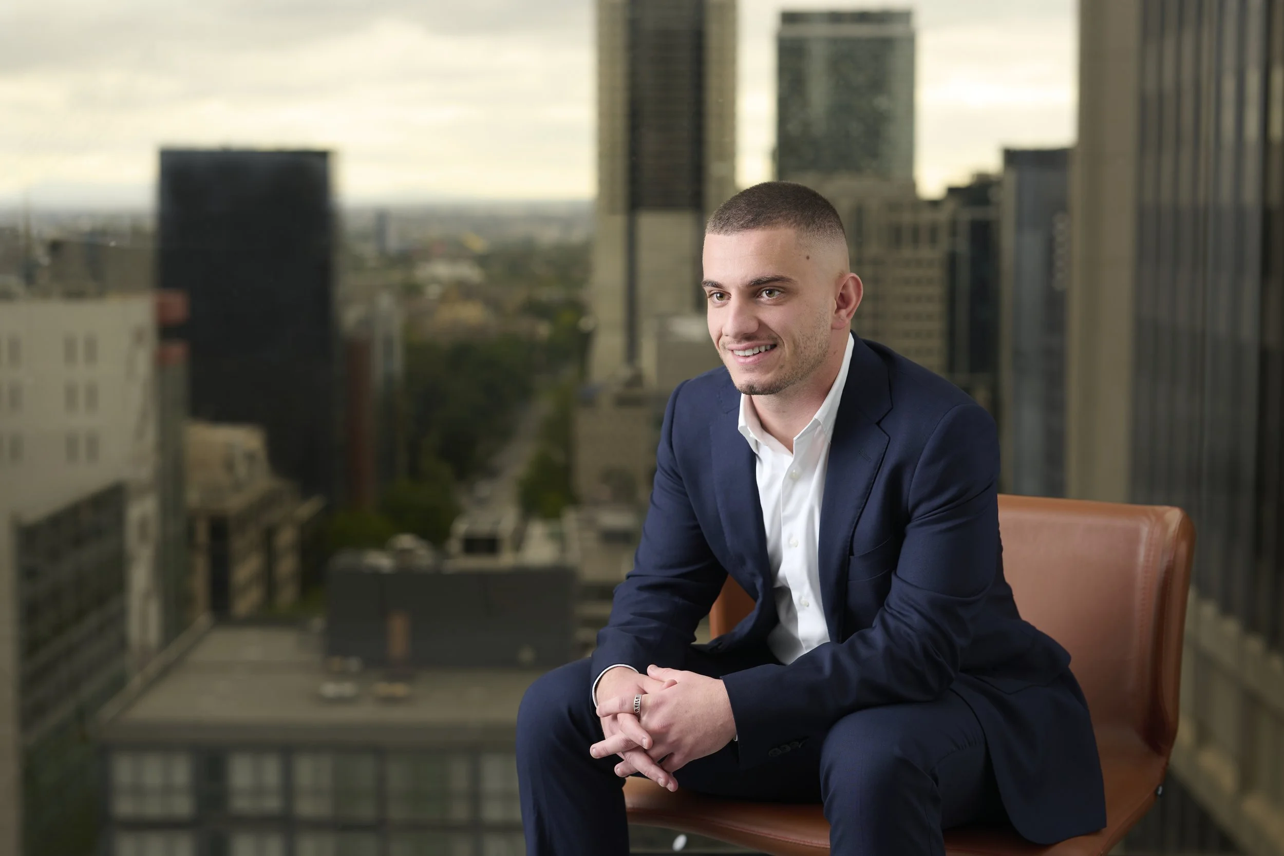 Smiling man in a navy suit seated in a leather chair with a city skyline visible through the windows