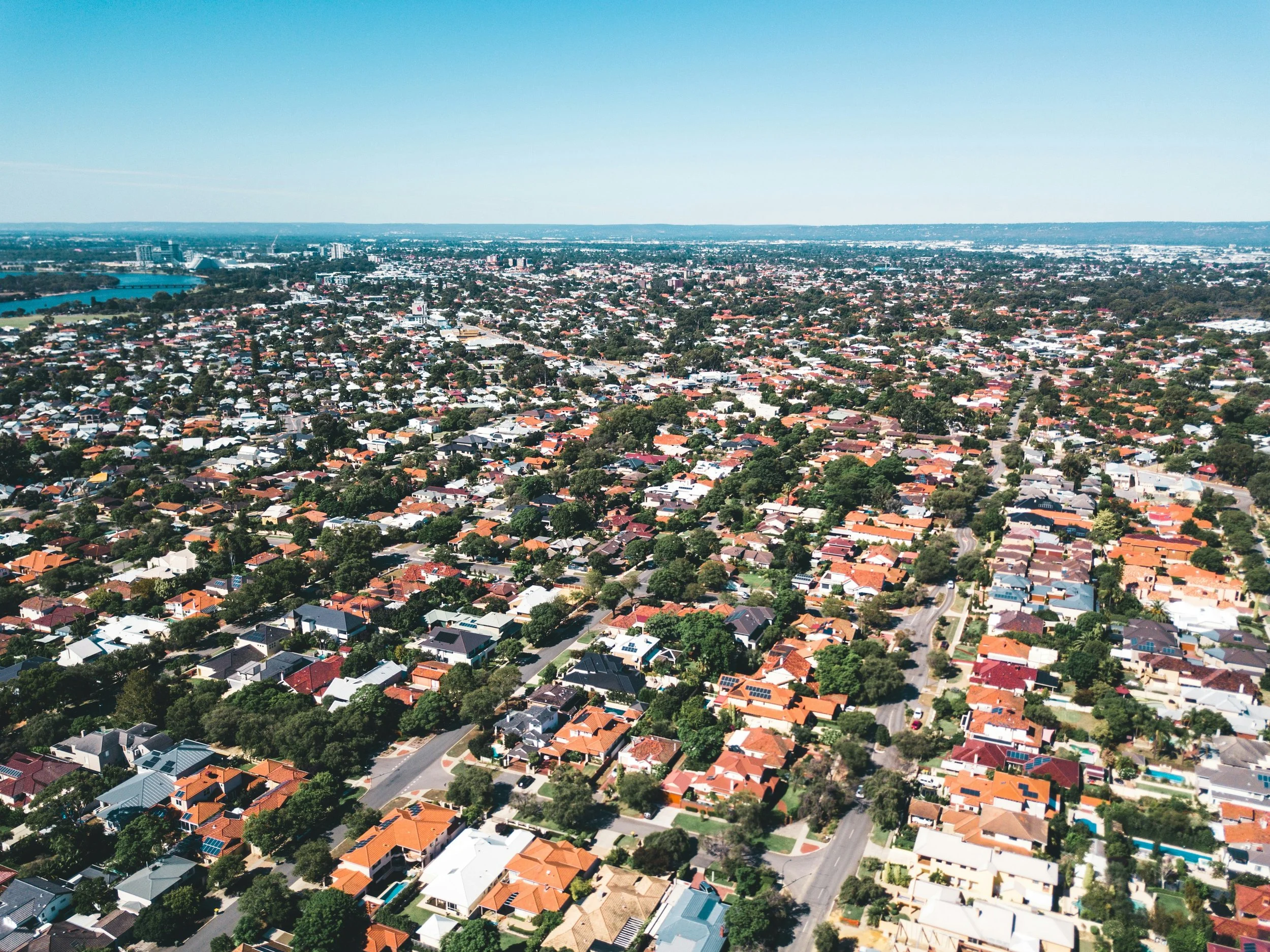 Aerial view of a sprawling residential cityscape with red-tiled roofs, tree-lined streets, a turquoise lake, and a blue horizon.