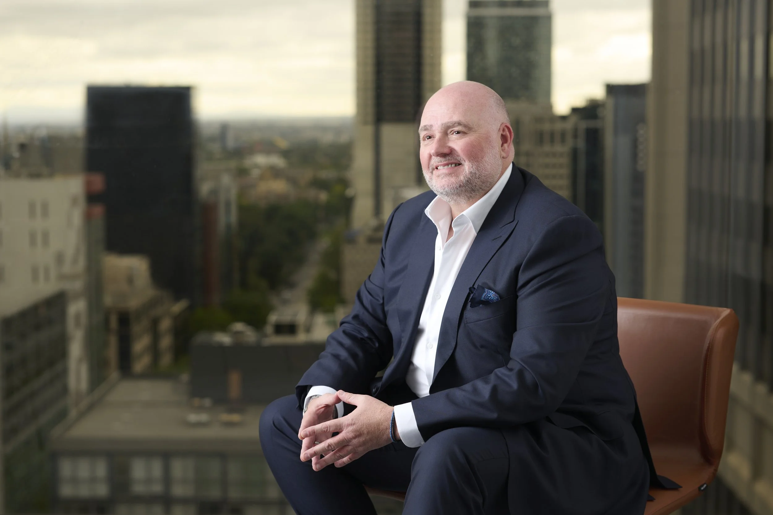 Bald businessman in a navy suit seated in a modern office overlooking a city skyline