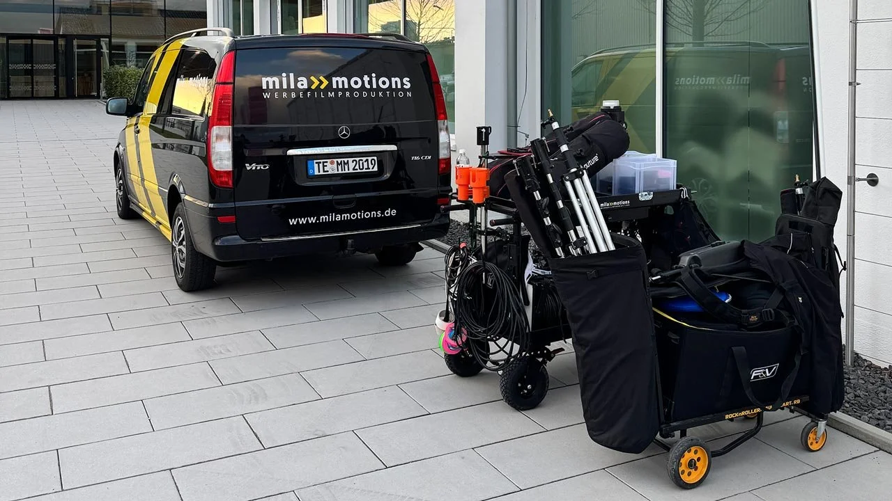 Ein van mit der Aufschrift "mila motions Werbefilmproduktion" und einer Ausrüstungskarren mit Kamera- und Filmausrüstung auf einem gepflasterten Boden vor einem modernen Gebäude. mila motions Werbefilmproduktion