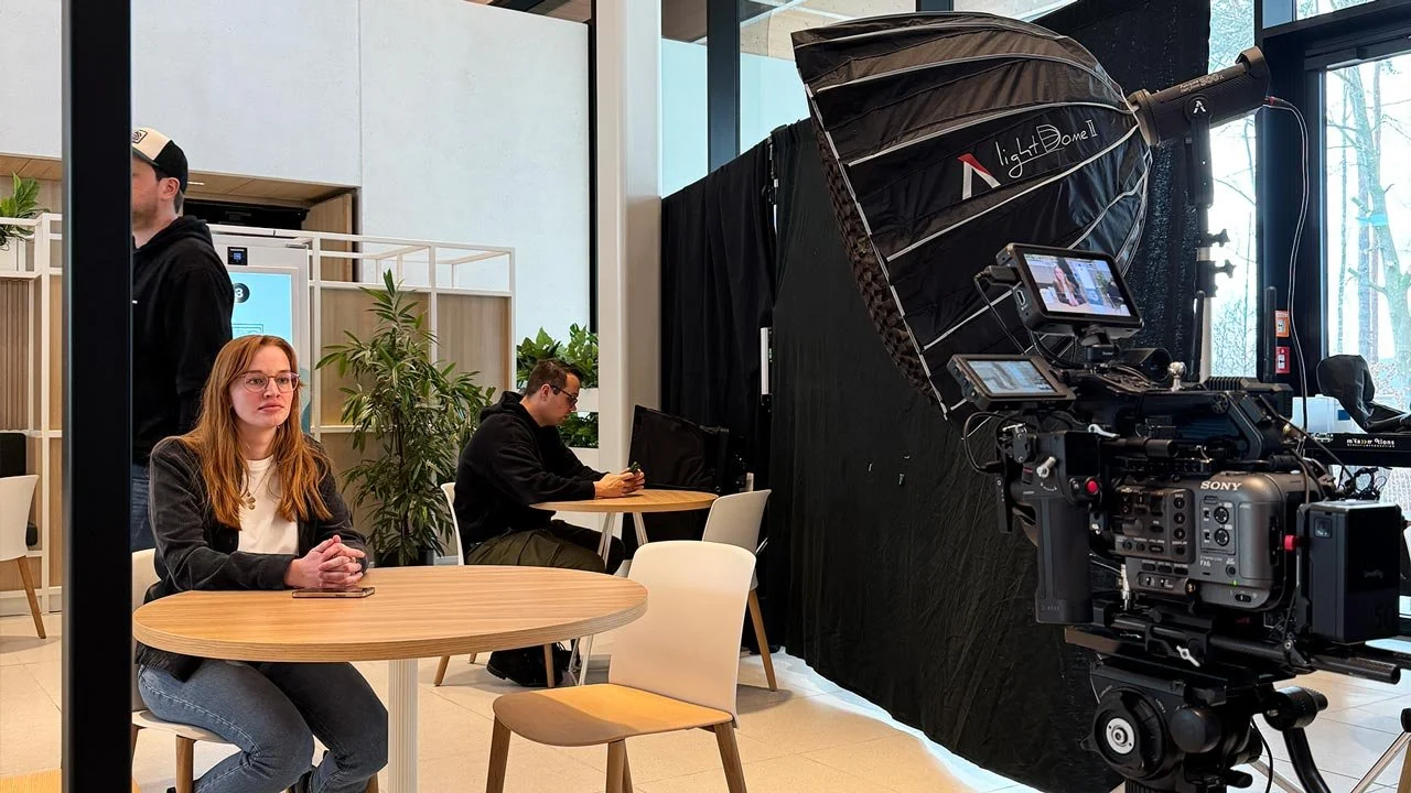 Aufnahme eines Videodrehs in einem modernen Raum mit Kamera, Lichttechnik, einem weißen Vorhang und mehreren Personen, darunter eine Frau mit roten Haaren, die an einem Tisch sitzt, und einen Mann, der an einem Laptop arbeitet. mila motions Werbefilm