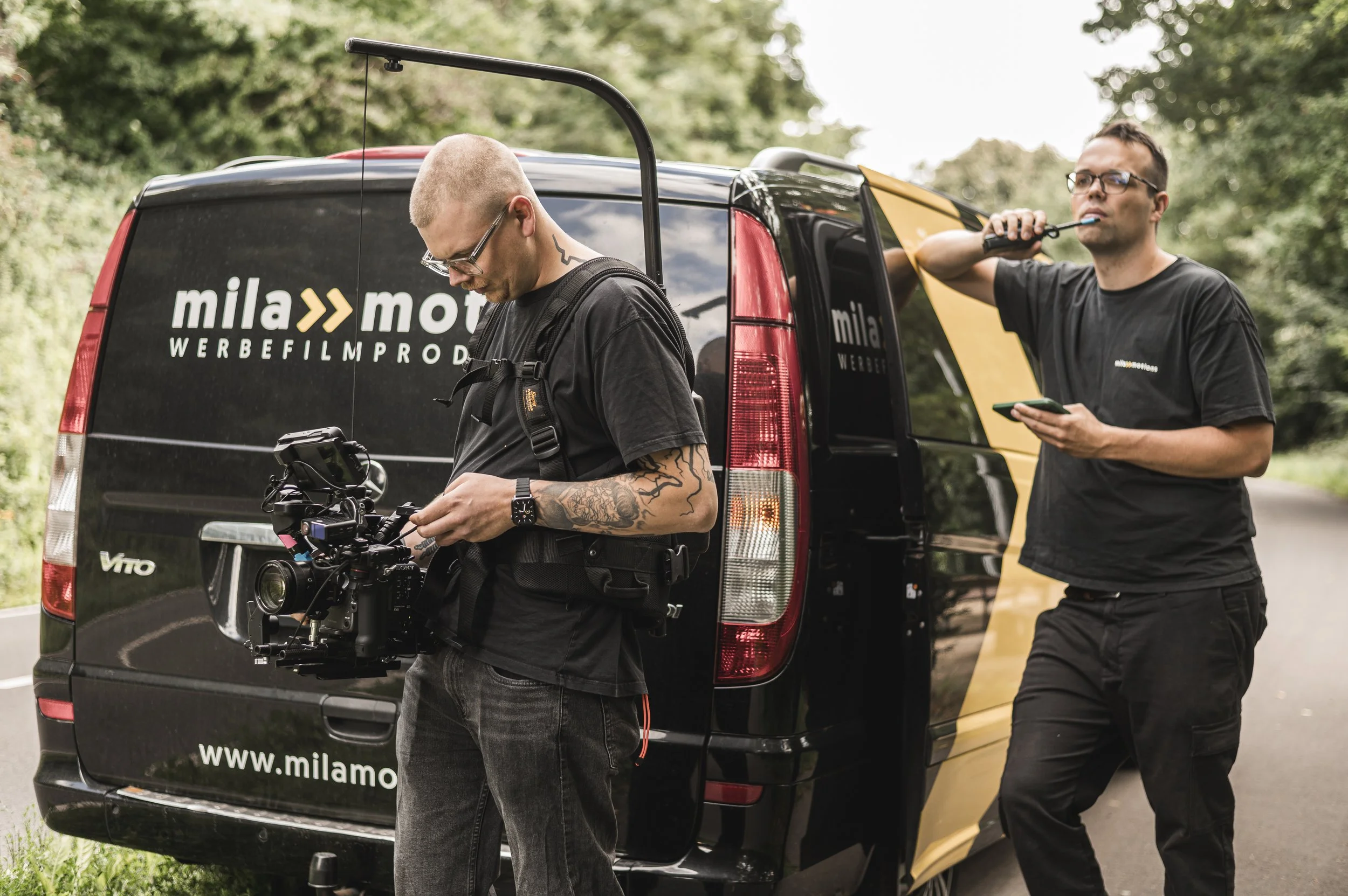 Zwei Männer arbeiten an einem schwarz-gelben Van, der das Logo "mila motions" und die Website "www.milamotions.de" trägt. Einer der Männer, mit Tattoos und Brille bedient eine Kamera. mila motions Werbefilmproduktion