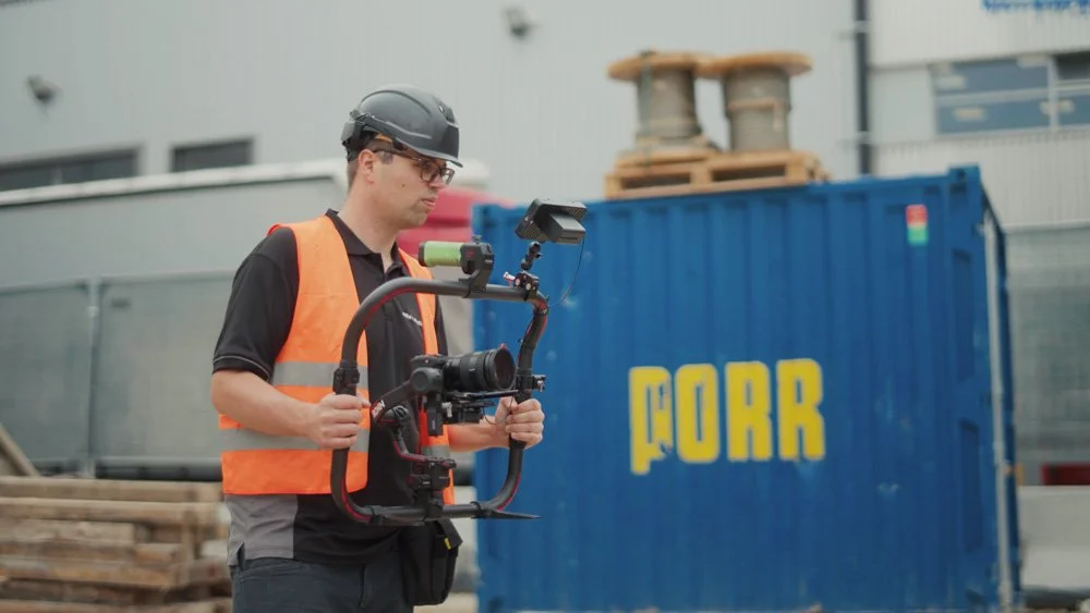 Ein Mann in Sicherheitsausrüstung mit Helm und Weste filmt mit einer Kamera auf einer Bauwand mit der Aufschrift 'PORR'. mila motions Werbefilmproduktion