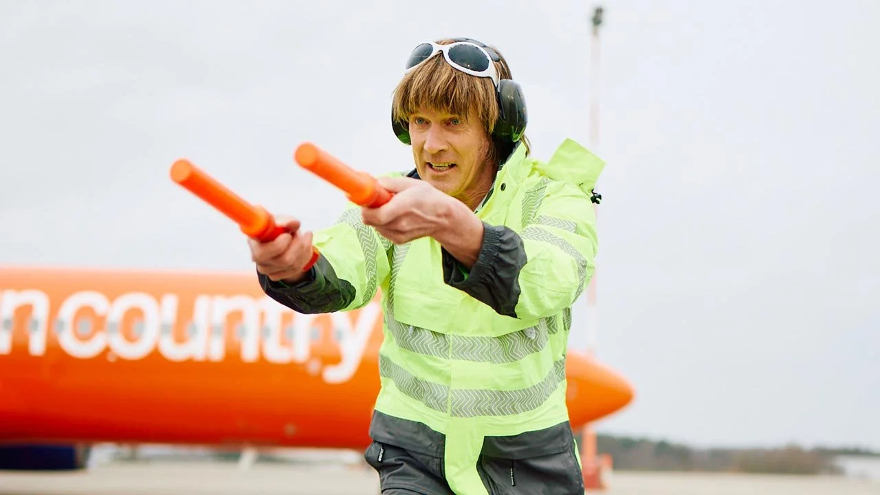 Mickie Krause mit Warnweste, Kopfhörer und Sonnenbrille zeigt mit beiden Händen rote Lichtwellenstäbe. mila motions Werbefilmproduktion