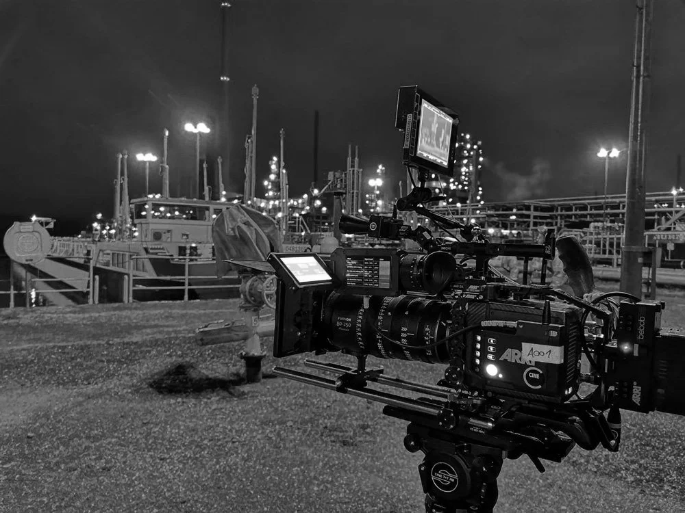 Filmkamera auf einem Stativ vor einem Hafen bei Nacht, mit Booten und Lichter im Hintergrund. mila motions Werbefilmproduktion