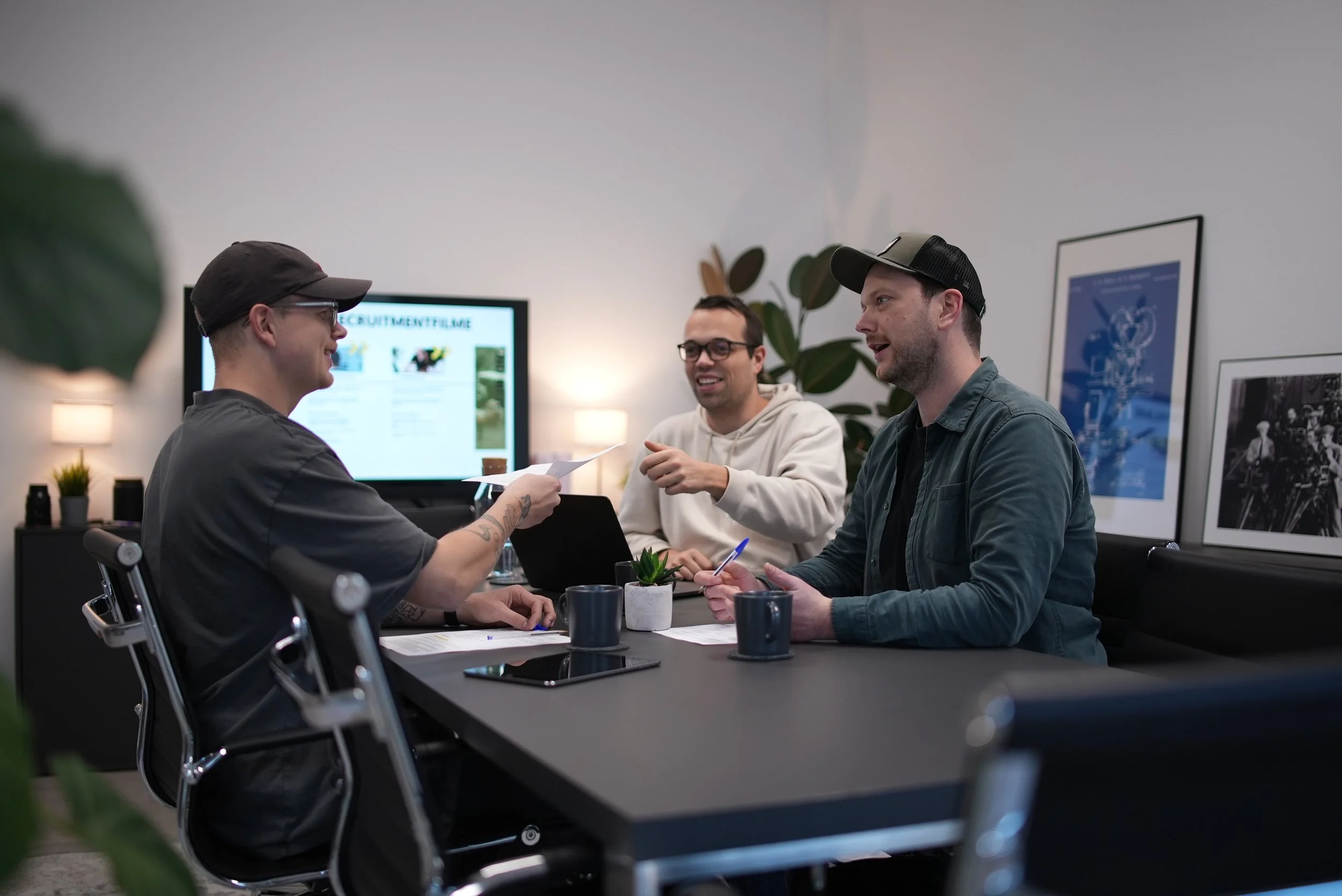 Drei Männer sitzen bei einem Meeting in einem modernen Büro, lachen und unterhalten sich, während sie Dokumente und Getränke auf dem Tisch haben. mila motions Werbefilmproduktion