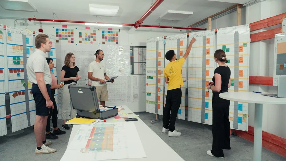 Menschen diskutieren und präsentieren in einem Raum mit großen Planern und Post-its an den Wänden, in einer Projektmanagement- oder Planungsbesprechung. mila motions Werbefilmproduktion