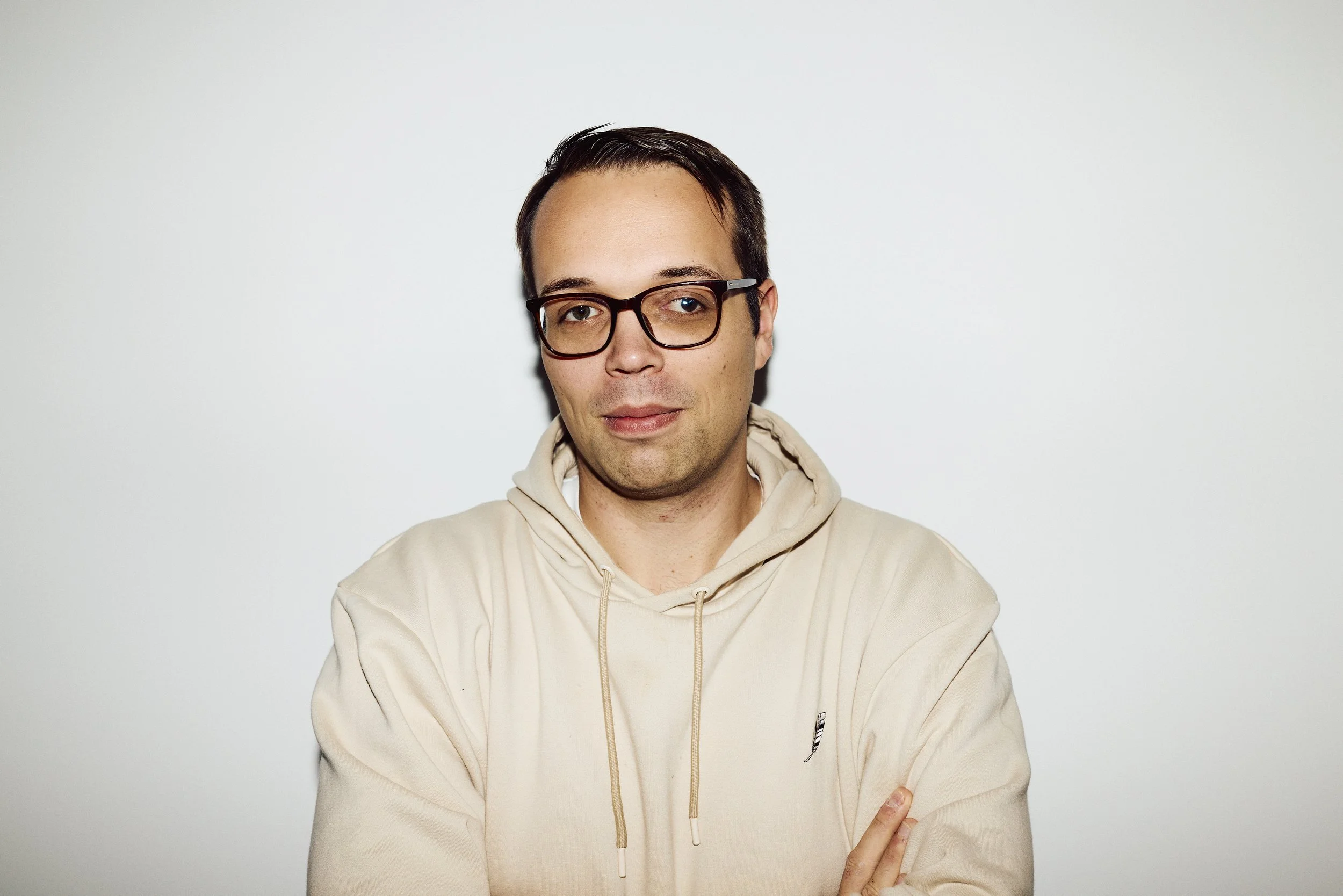 Junge Mann mit dunklen Haaren, Brille und beigem Hoodie steht vor weißem Hintergrund. mila motions Werbefilmproduktion