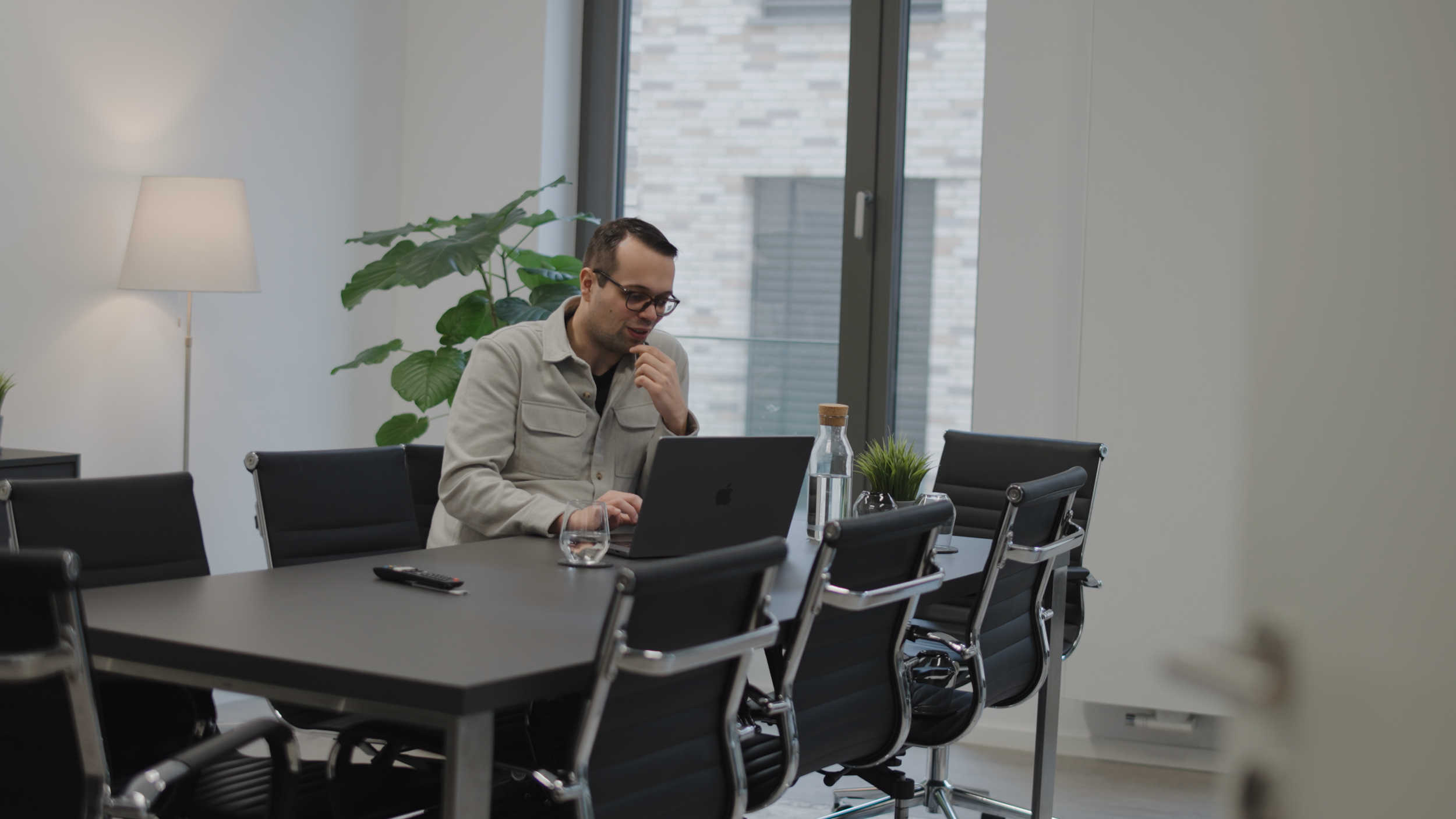 Ein Mann sitzt in einem modernen Besprechungsraum vor einem Laptop und schaut auf den Bildschirm, während er nachdenklich aussieht. Der Raum ist minimalistisch eingerichtet mit dunklen Stühlen, einer Tischplatte, einer Wasserflasche, einem Glas, einer Pflanze und einem Stehlampe neben einem großen Fenster.