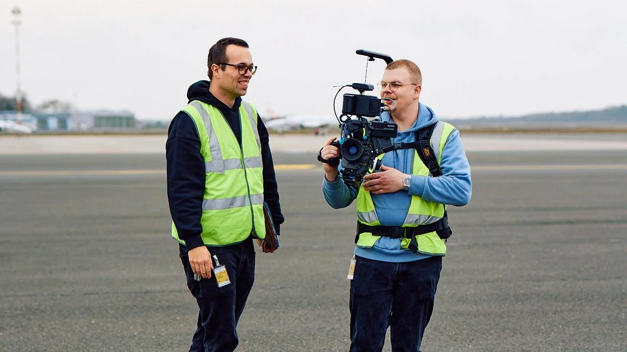 Zwei Männer in Hochsichtwesten, die auf einem Flugplatz stehen, einer hält eine Kamera auf Schulterhöhe. mila motions Werbefilmproduktion
