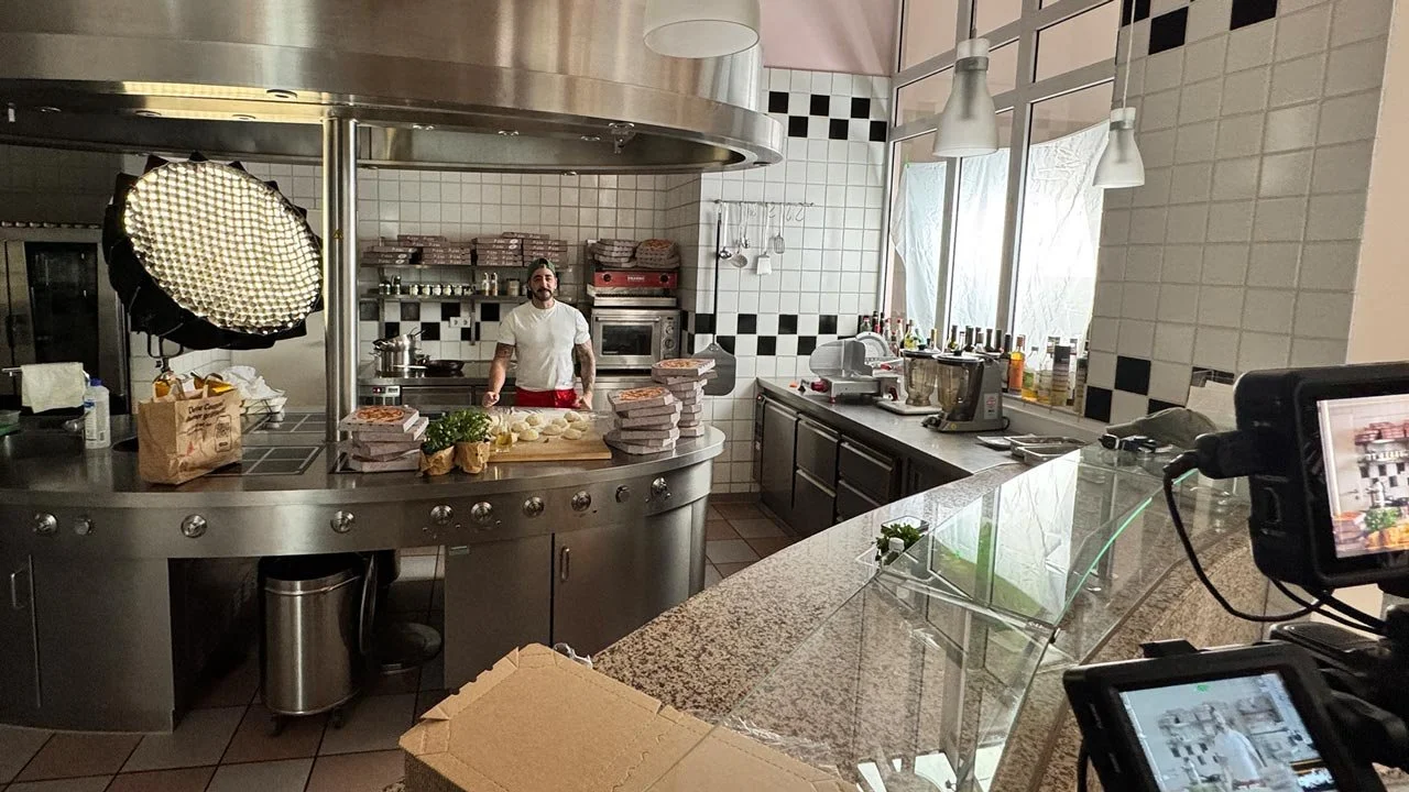 Ein professionelles Küchenstudio mit einem Koch, der vor einem Kochfeld steht, umgeben von Kuchen und Zutaten. Es gibt Küchengeräte, Flaschen, und große Fenster bringen viel Licht in die Küche. mila motions Werbefilmproduktion