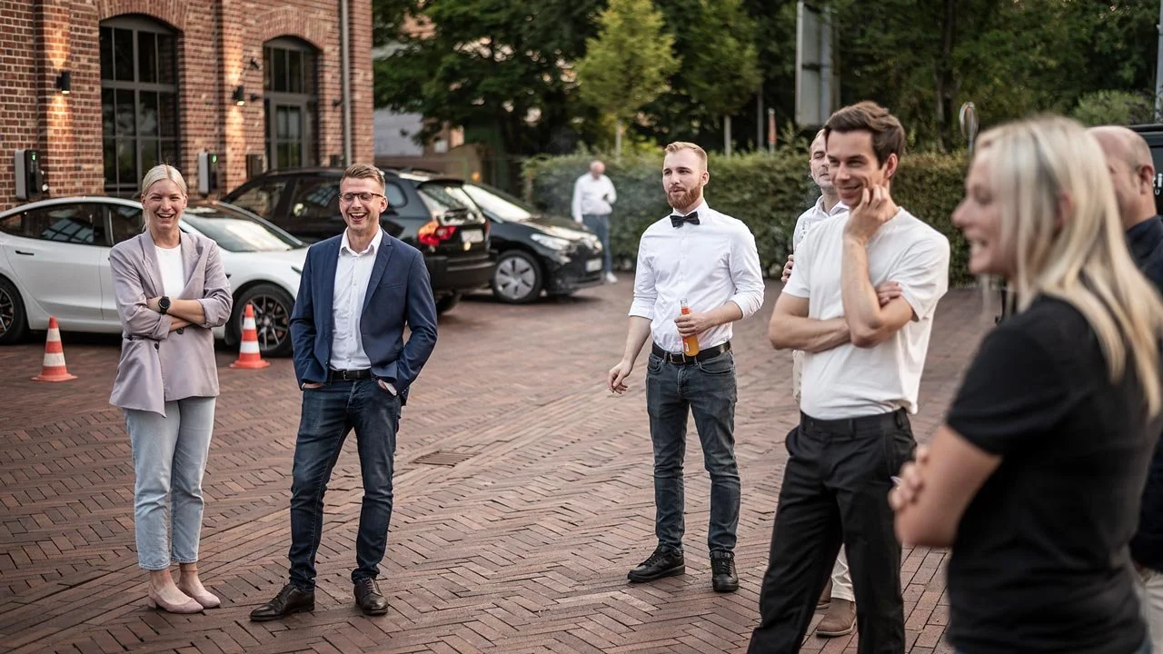 Eine Gruppe von Menschen, die im Freien stehen und lachen,mit Autos im Hintergrund auf einer gepflasterten Straße. mila motions Werbefilmproduktion