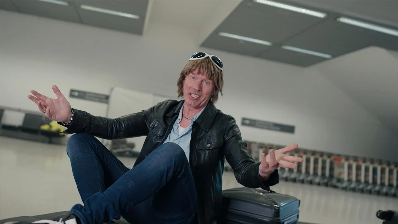 Ein Mann mit braunen, mittellangen Haaren trägt eine Lederjacke und trägt eine schwarze Tasche. Er sitzt in einem Flughafen und wirkt fröhlich, während er mit den Armen gestikuliert. mila motions Werbefilmproduktion