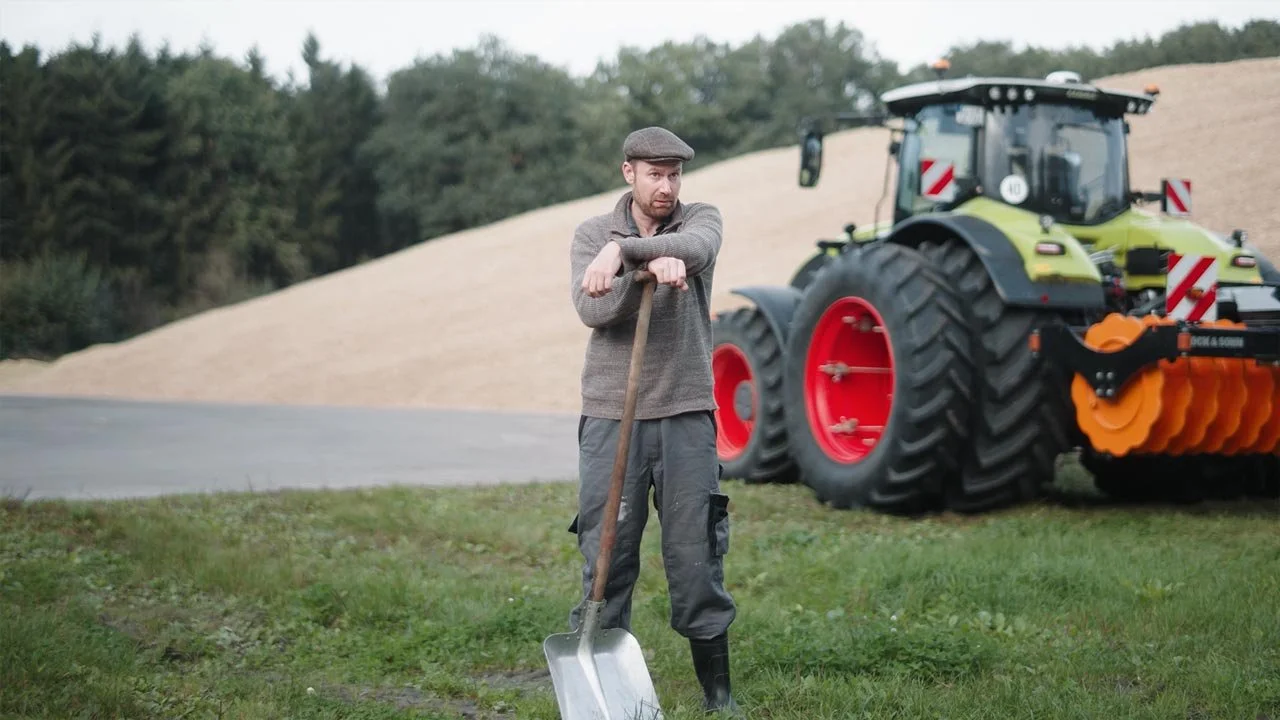 Ein Mann lehnt auf einem Spaten, im Hintergrund ein landwirtschaftlicher Traktor auf grünem Feld. mila motions Werbefilmproduktion