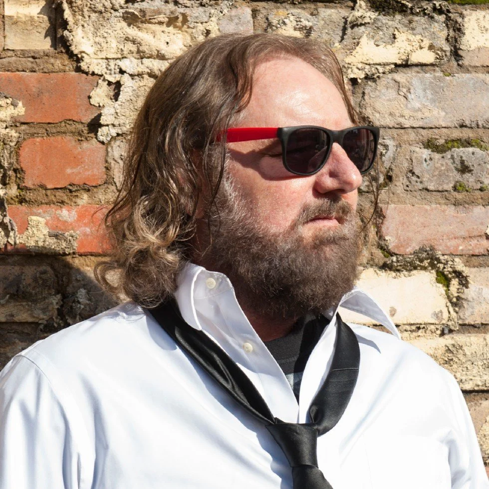 Brett Mitchell headshot in sunglasses next to brick wall