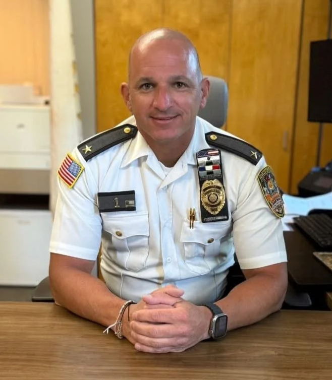 Chief Jeffrey M. Jenison sitting in a chair at a light brown wooden desk in his Police uniform
