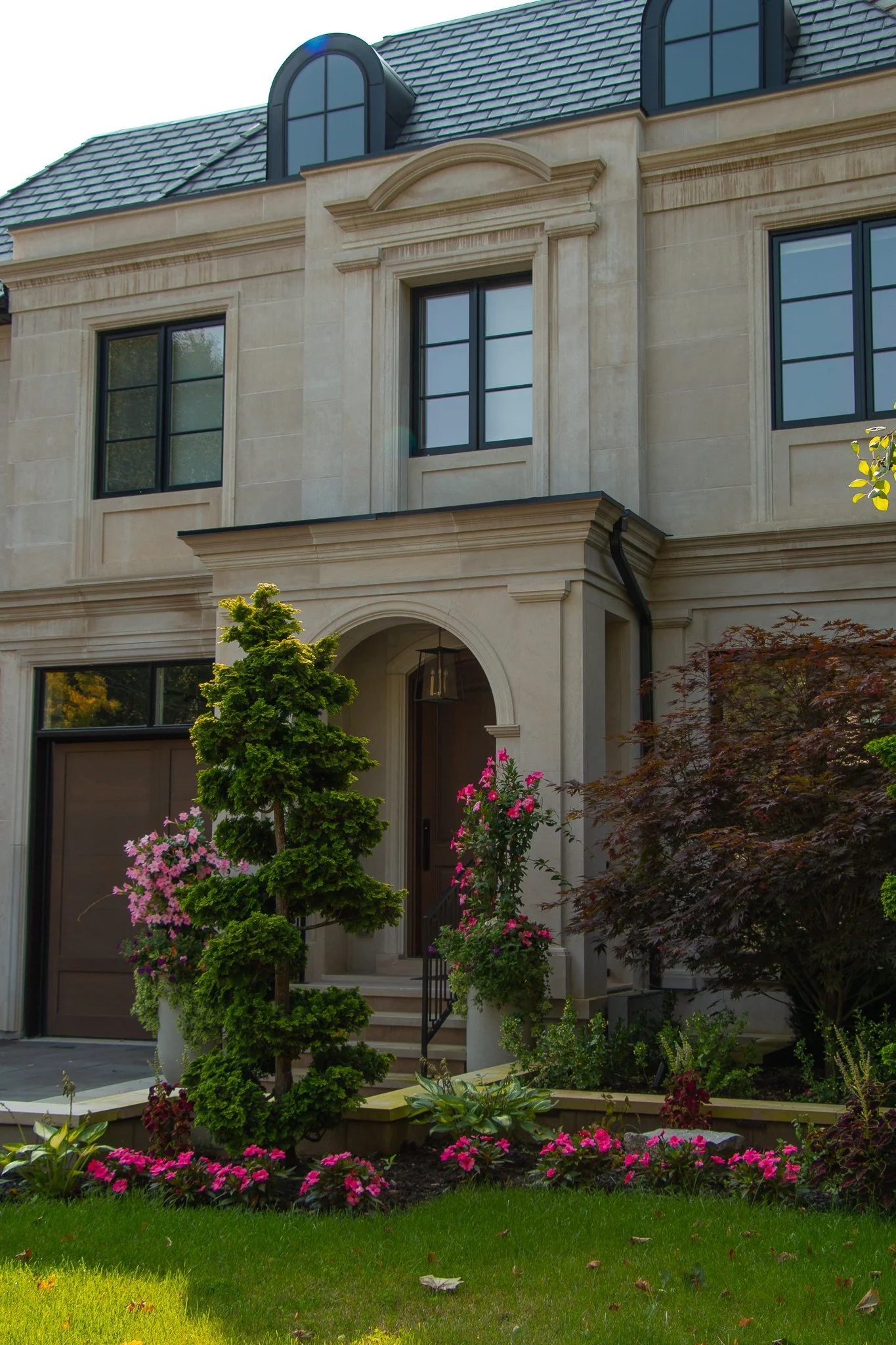 Front view of an elegant house with large windows, a small garden with a manicured tree, pink flowers, and green shrubbery.