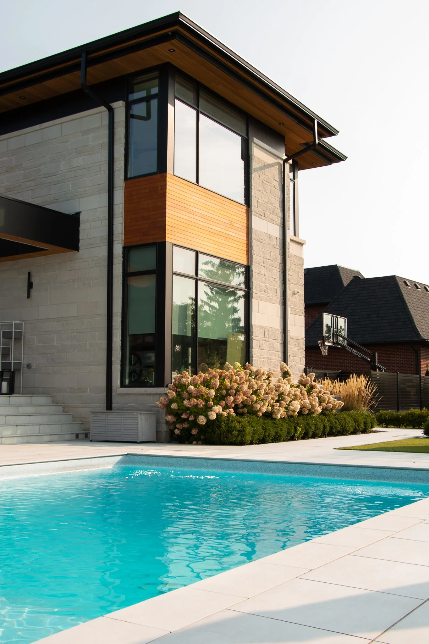 Modern house with large windows, stone and wooden exterior, surrounding landscaped yard with blooming hydrangeas, view of a pool in the foreground, and neighboring houses in the background.