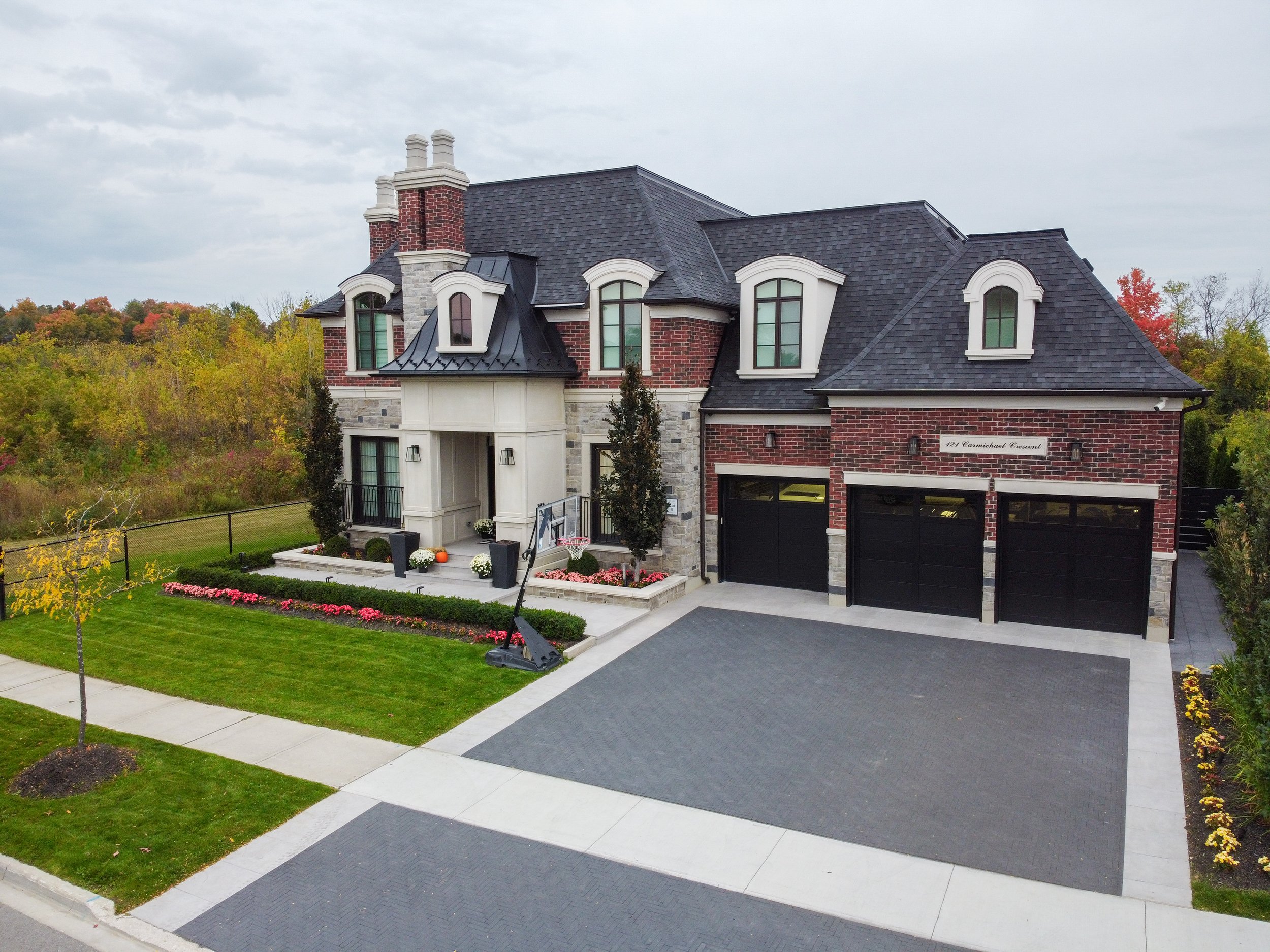 Modern two-story house with brick and stone exterior, black garage doors, driveway, and landscaped yard with grass, trees, and flowers.