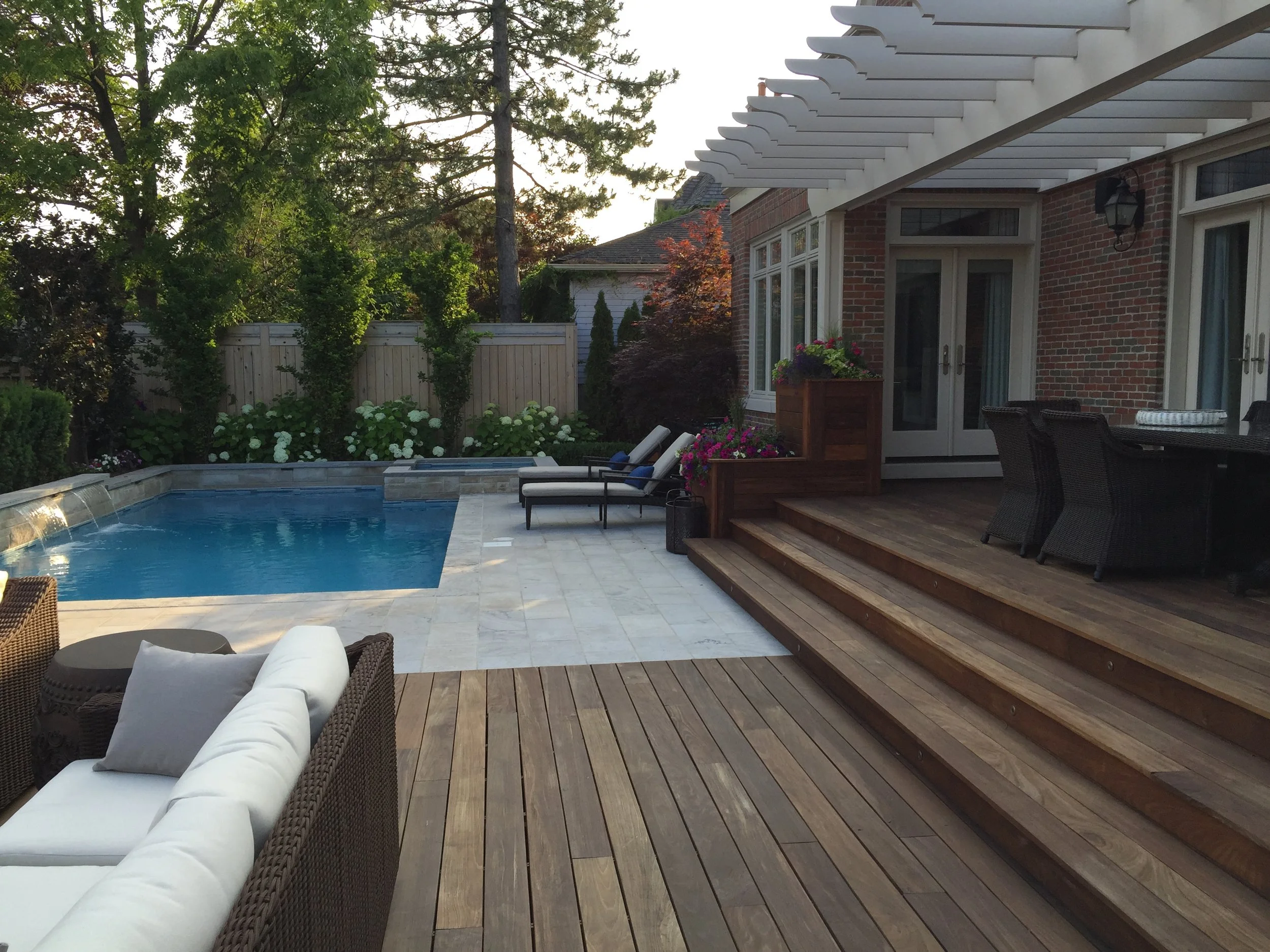 Backyard patio with swimming pool, lounge chairs, outdoor dining table, and potted flowers, surrounded by a wooden deck and a brick house with large windows and a white pergola.