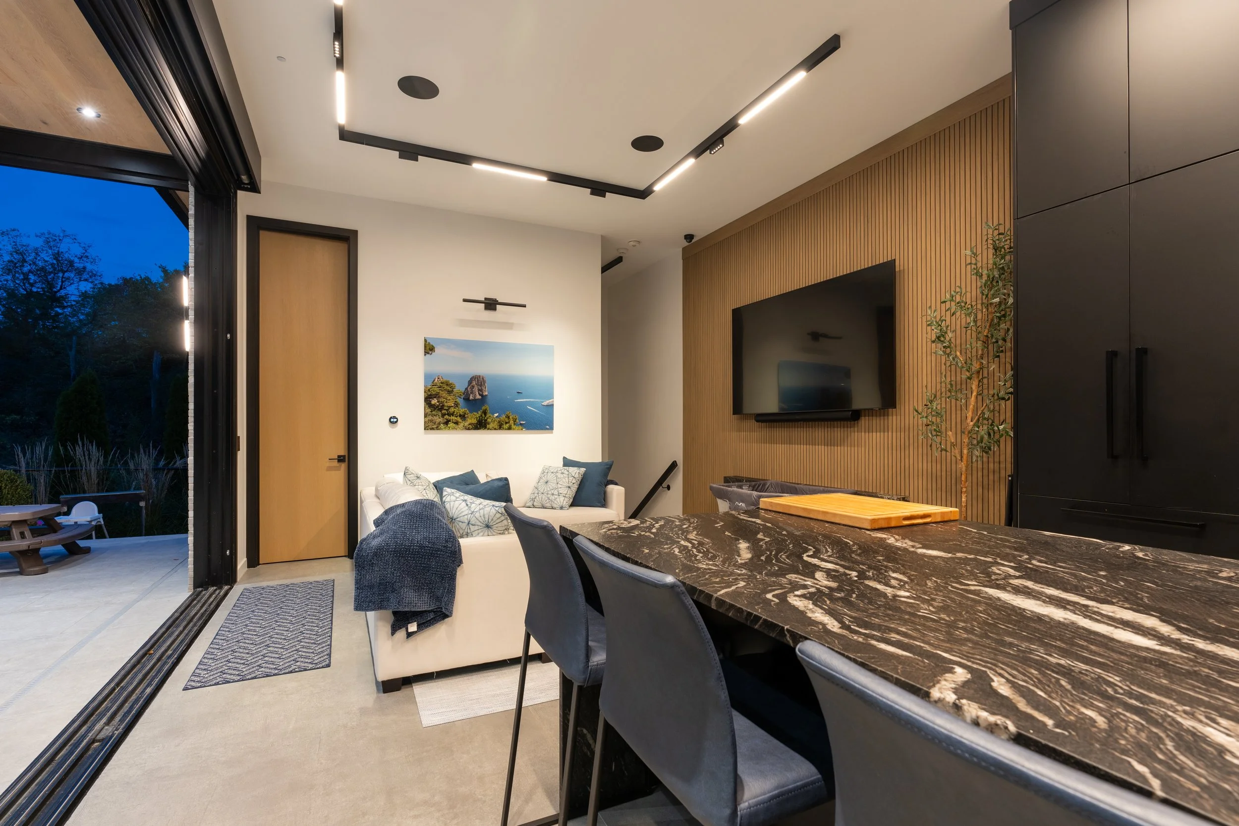 Modern living and dining room with a marble-topped table, black chairs, a beige sofa with blue cushions, a wall-mounted TV on a wooden accent wall, a large sliding door open to an outdoor patio, and contemporary ceiling lighting.