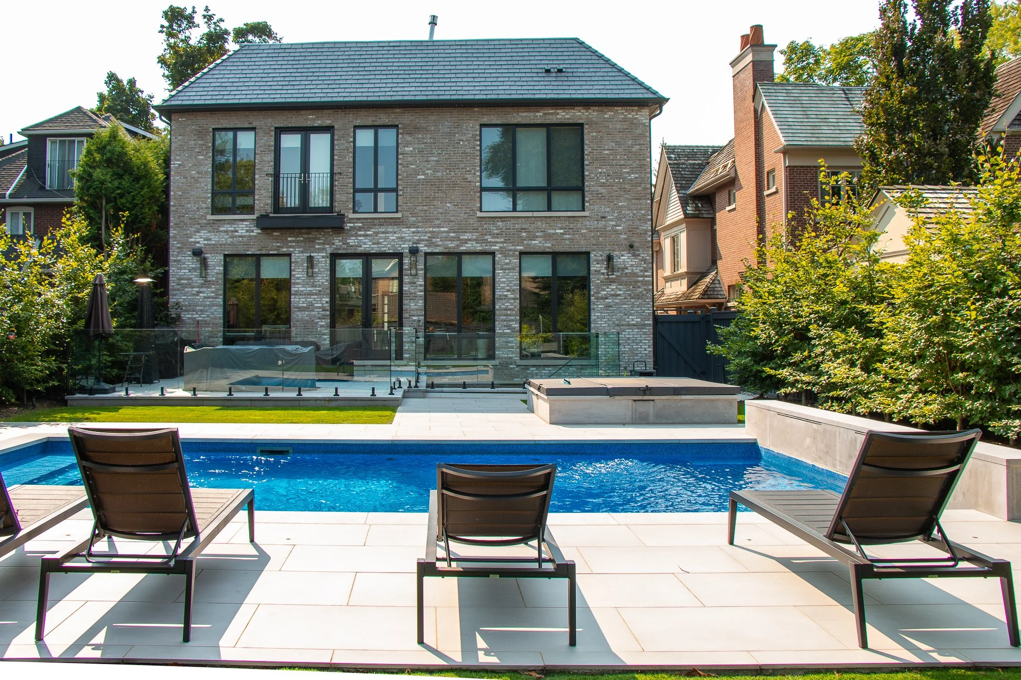 Backyard with rectangular swimming pool, three brown lounge chairs, green trees, modern brick house with large windows and a balcony in the background.
