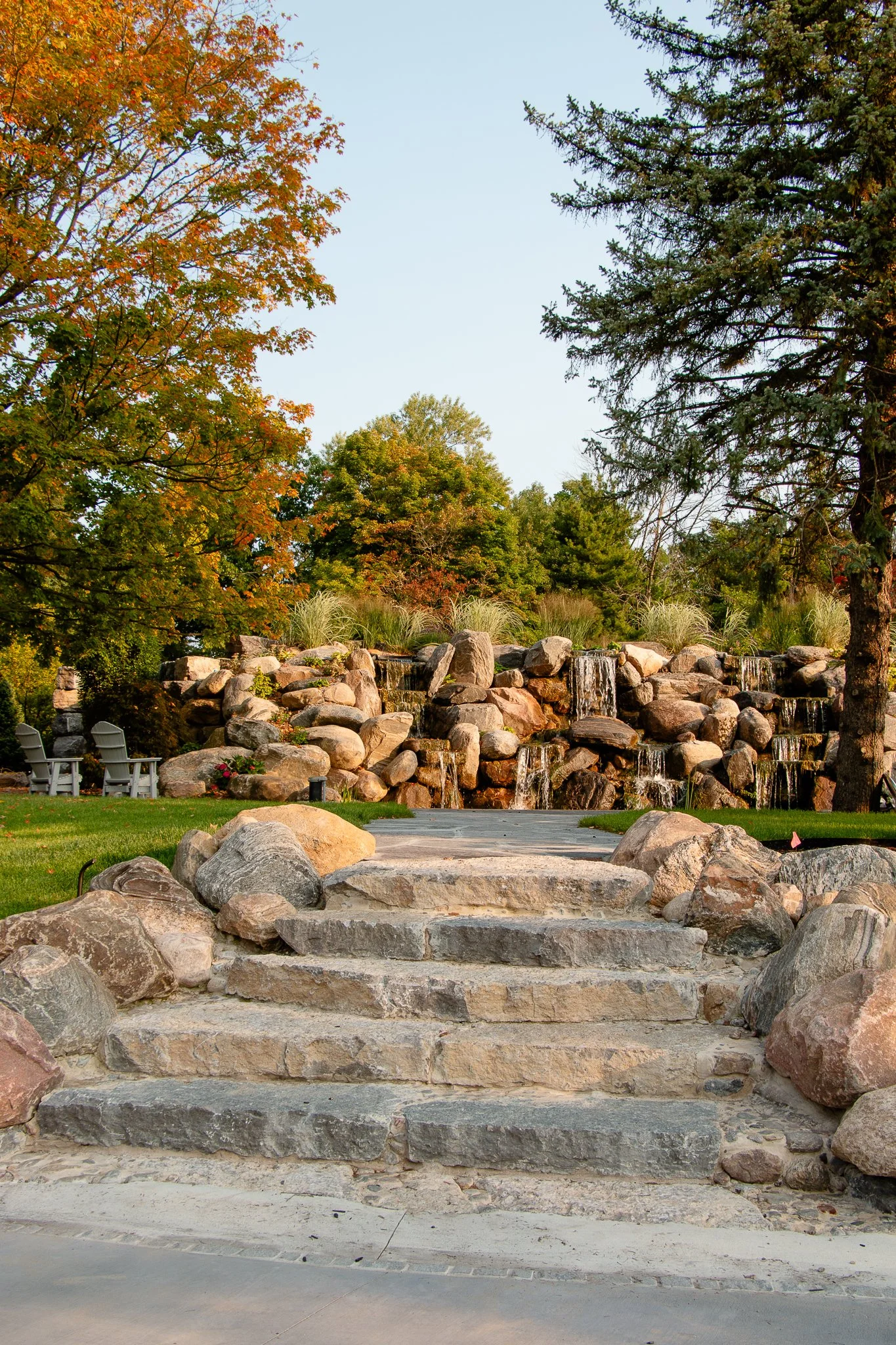 A stone staircase leading to a landscaped yard with a waterfall feature made of rocks, surrounded by trees with autumn foliage, two chairs on the grass, and a clear blue sky.