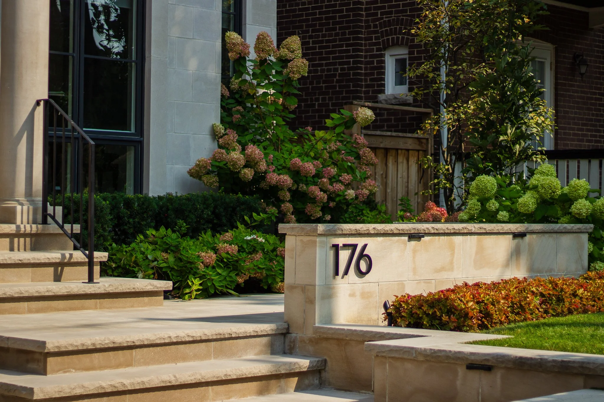 Front entrance to a residential building with stairs, a stone wall with the number 176, and landscaped bushes and flowers.