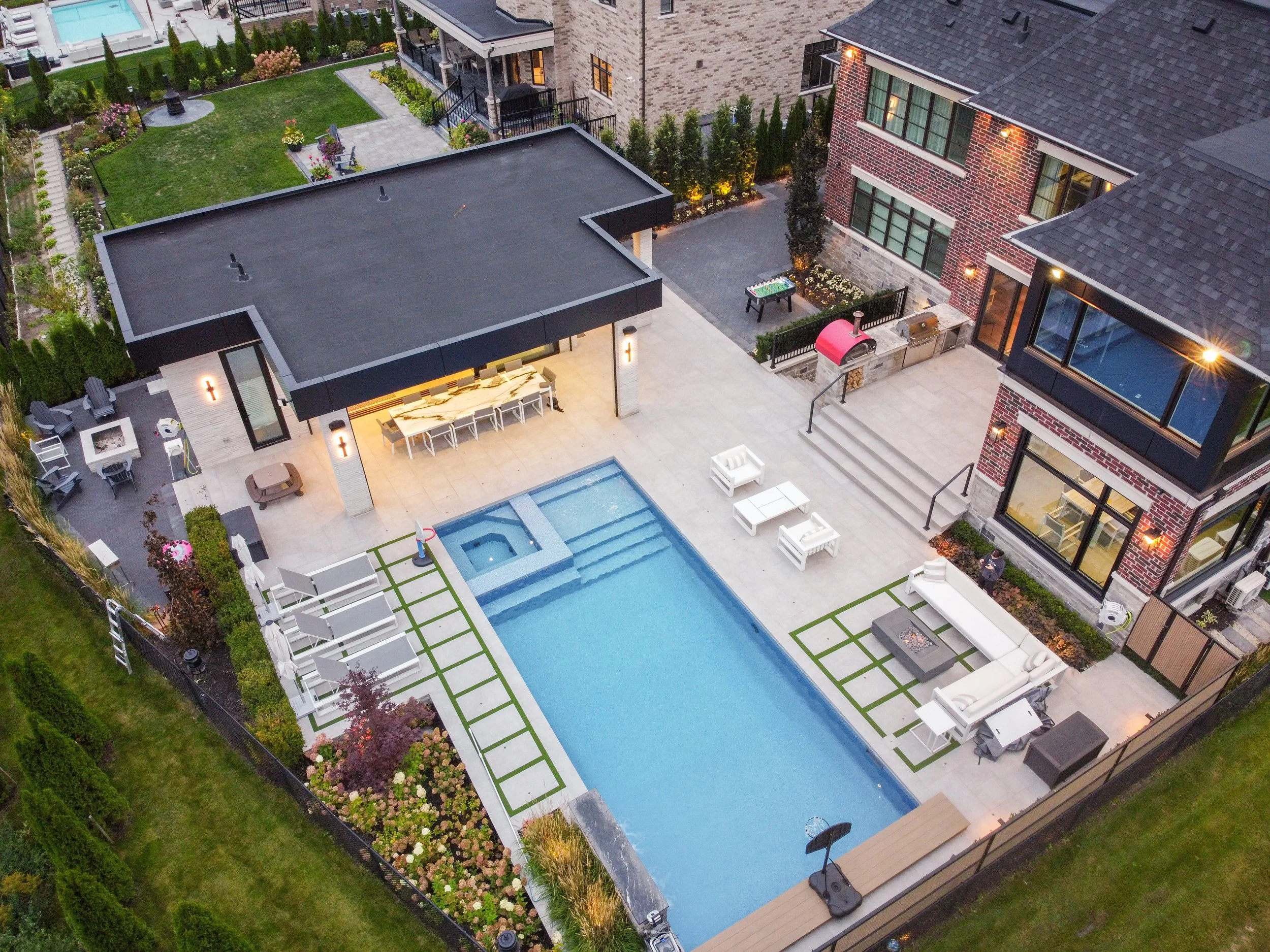 Aerial view of a modern residential backyard with a swimming pool, outdoor seating, fire pit, a covered patio area with an outdoor kitchen, landscaped garden, and neighboring houses.