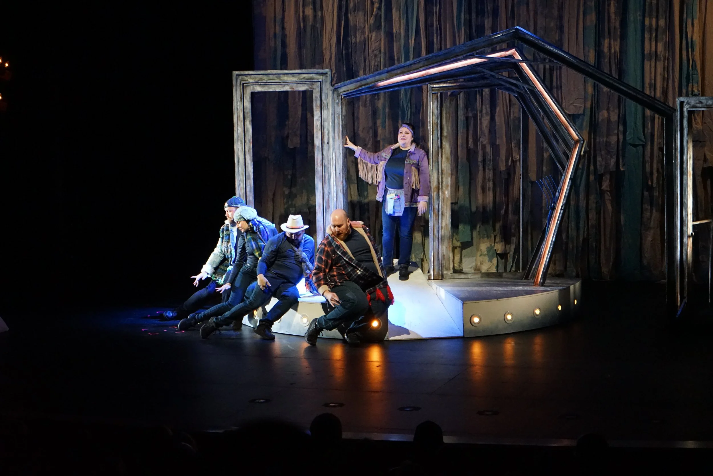 Une scène de théâtre avec plusieurs acteurs en costume, certains sont assis ou accroupis, un est debout avec les bras tendus, une femme se tient sur une plateforme en arrière-plan, avec un décor de bois derrière eux.